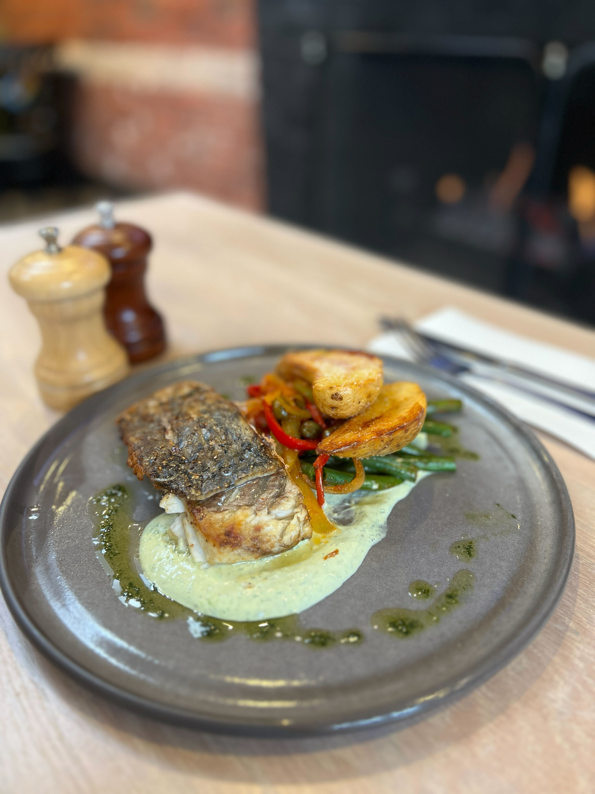 A plate of food with a piece of cooked fish, topped with green vegetables, colorful peppers, and potato wedges, on a dark round plate.