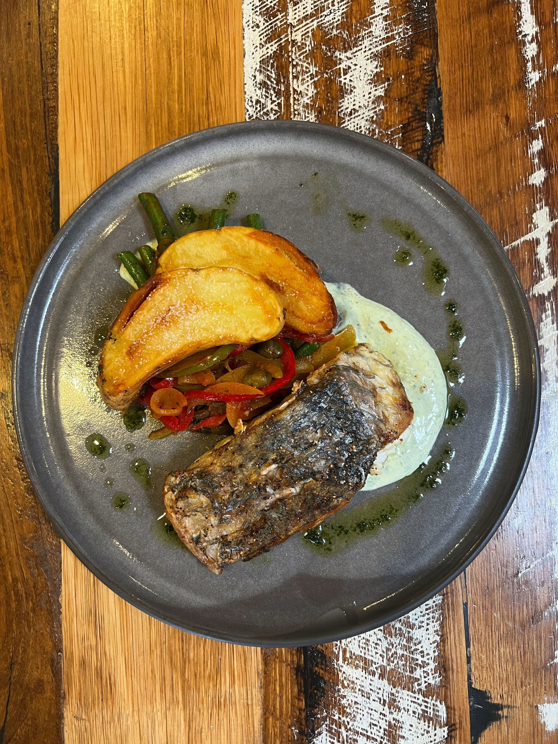 A plate of grilled fish served with potato wedges, sautéed peppers and green beans, garnished with green sauce and a creamy white sauce, on a wooden table.