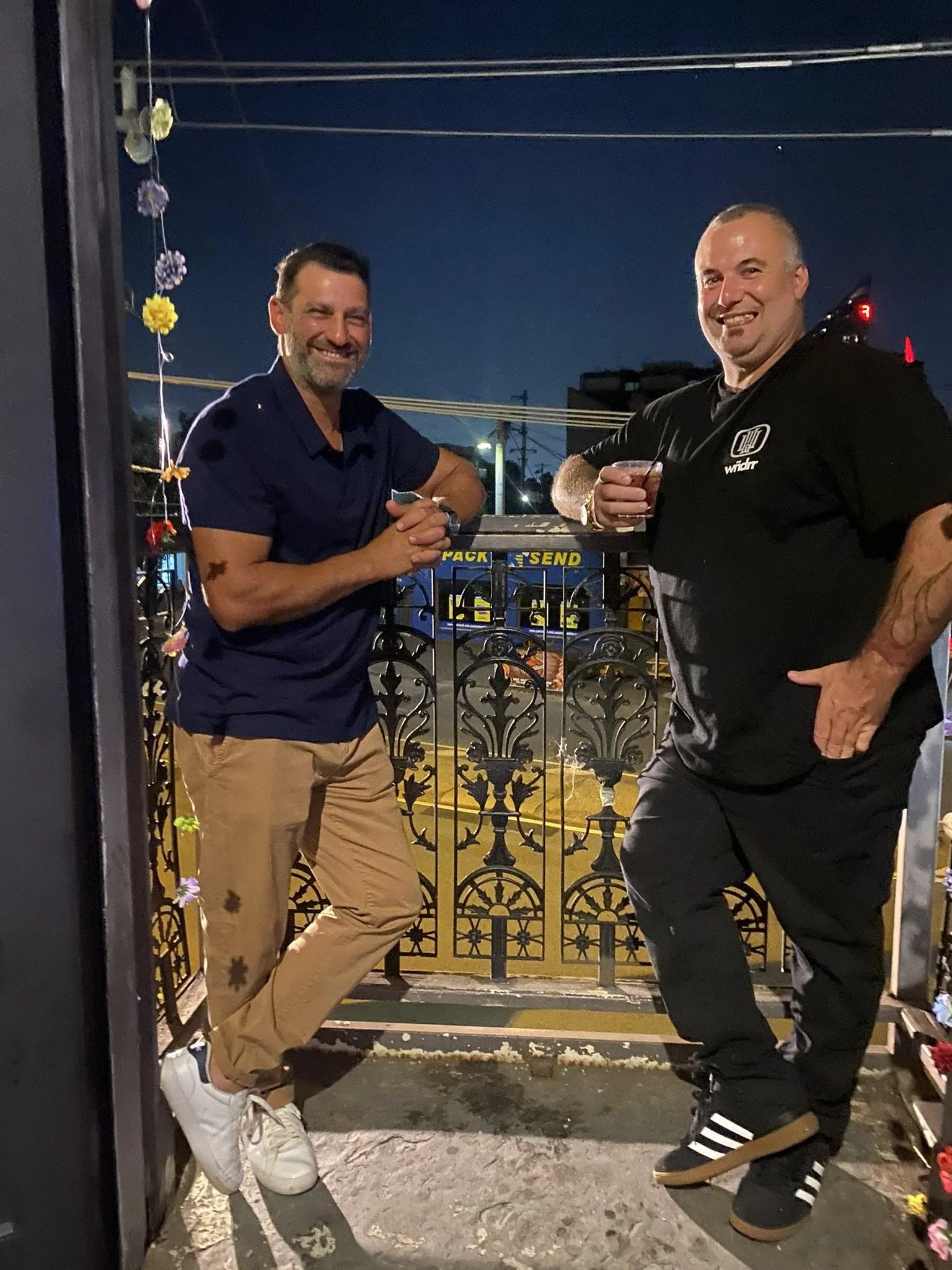 Two men smiling and posing on a balcony at night upstairs at the Palace Hotel. One is wearing a navy blue polo and beige pants, and the other is in a black T-shirt and dark pants, holding a drink. 