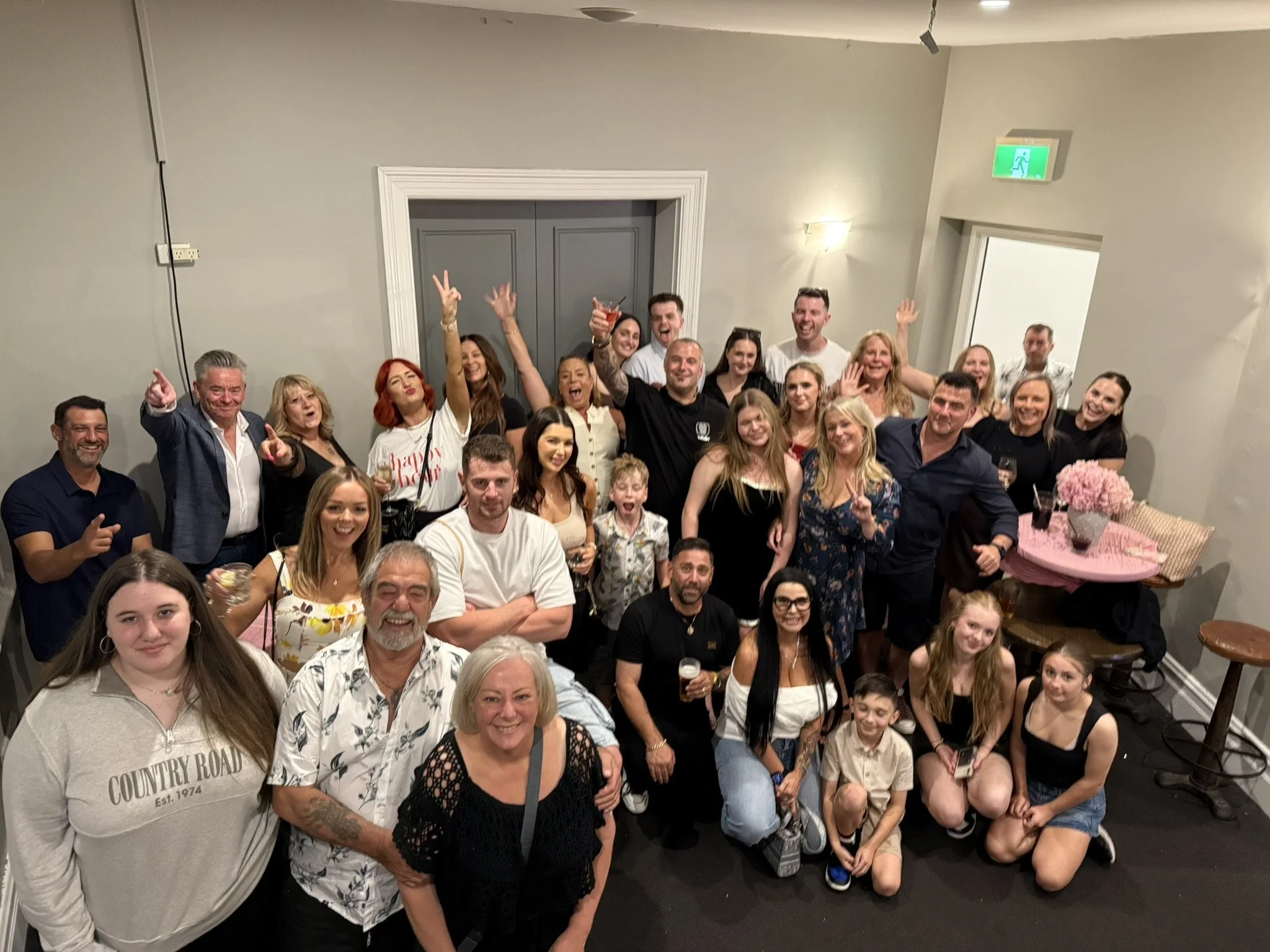 Group of smiling people at a celebration in a room, some holding drinks, posing for a group photo.