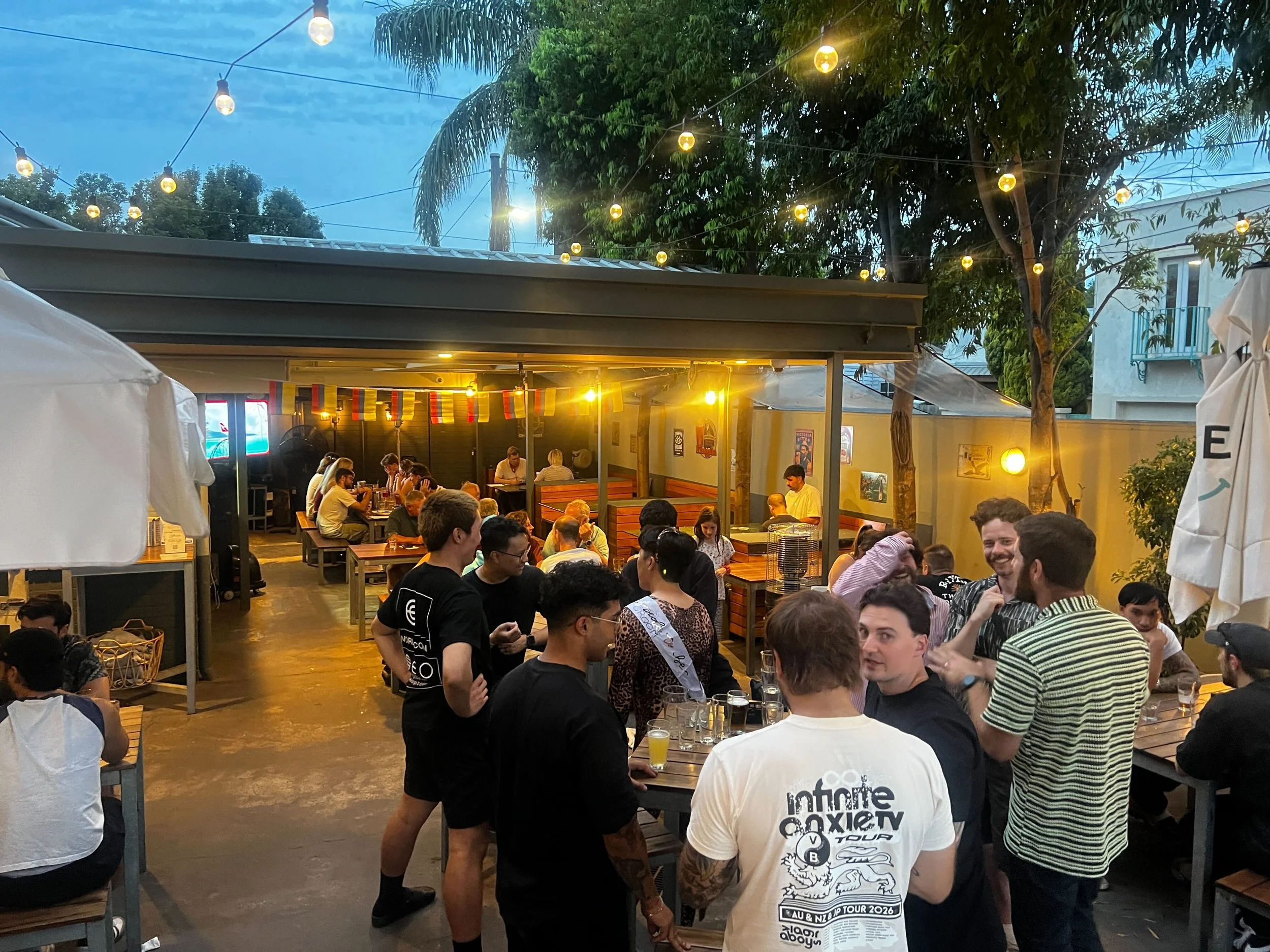 Outdoor gathering with people dining under string lights at a restaurant in the evening, surrounded by trees and buildings.