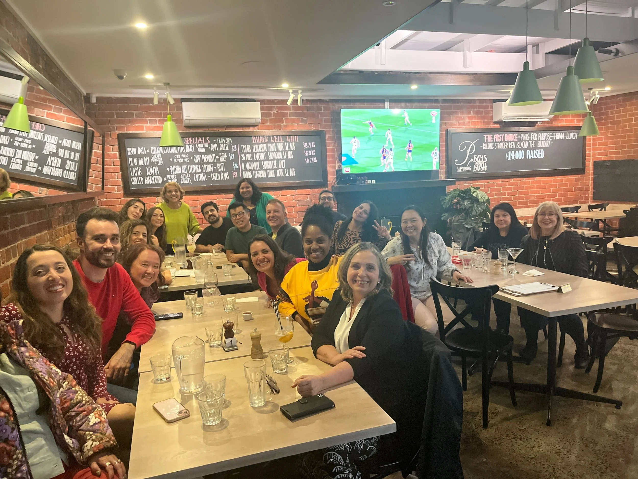 A large group of people sitting at a long dining table in a restaurant, smiling and enjoying a meal together.