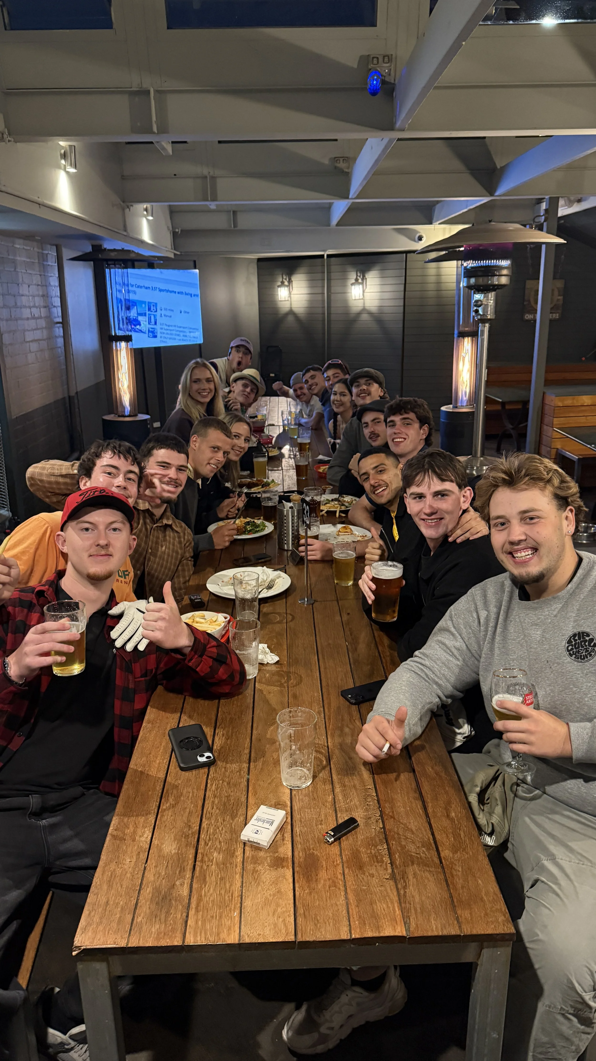 A group of young adults sitting around a long wooden table at a restaurant or bar, enjoying food and drinks, smiling and posing for the camera.