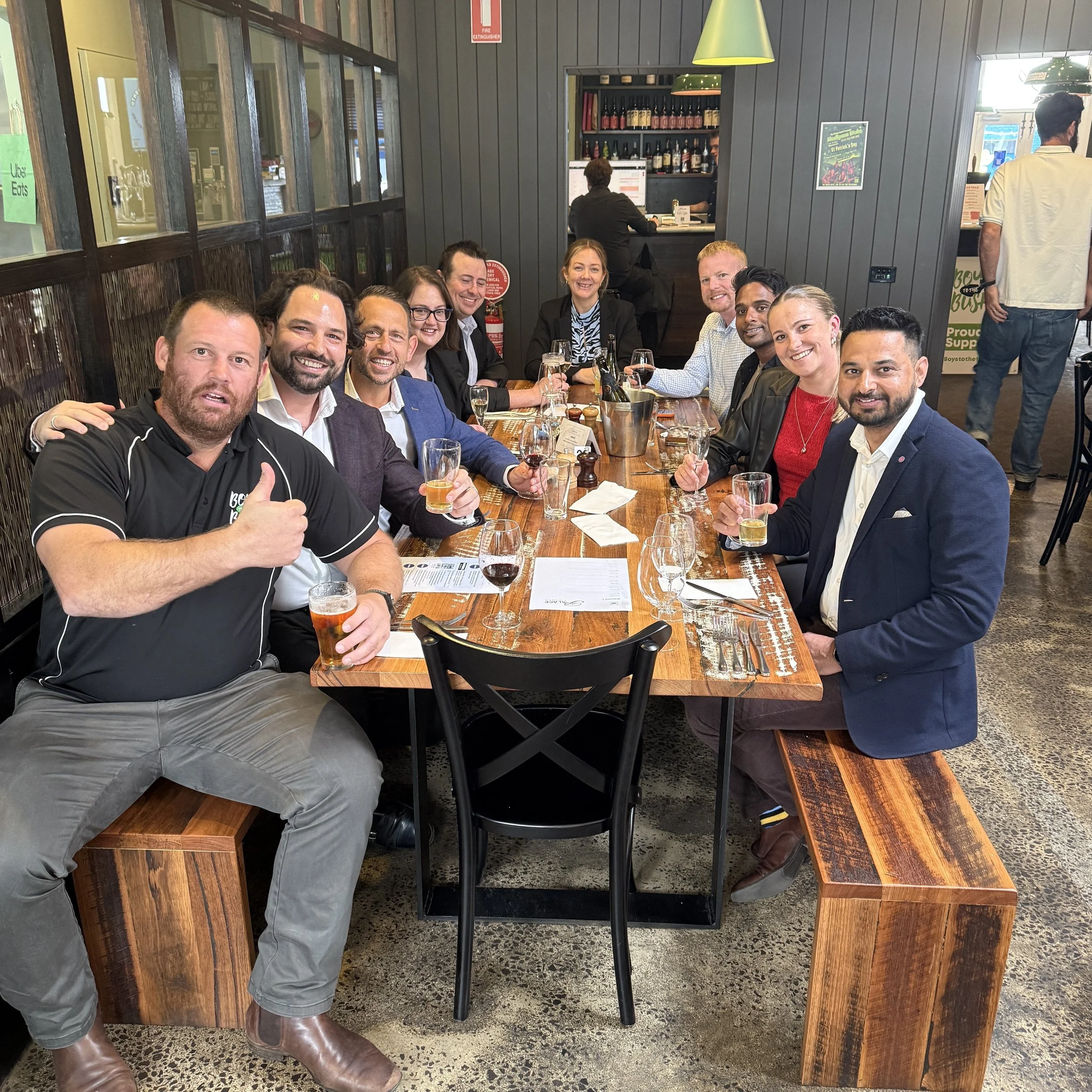 A group of people sitting around a wooden table in a restaurant, smiling and holding glasses of wine or beer. There are nine people visible, dressed in casual and semi-formal clothing. The background features a wall with shelving of bottles, a person