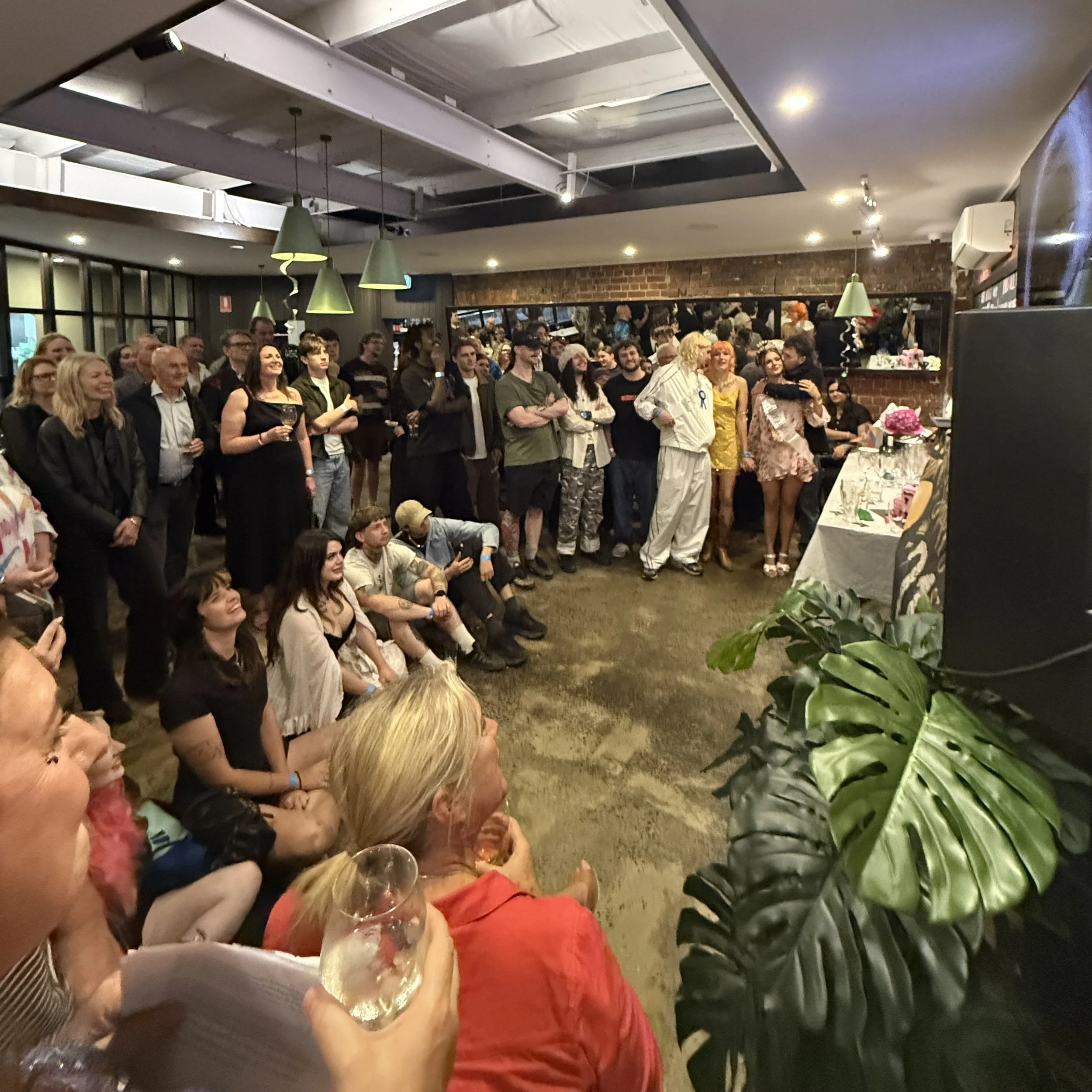A group of people attending a social event, some standing and some sitting, with a reception table in the background. The room has a brick wall and green pendant lights hanging from the ceiling.