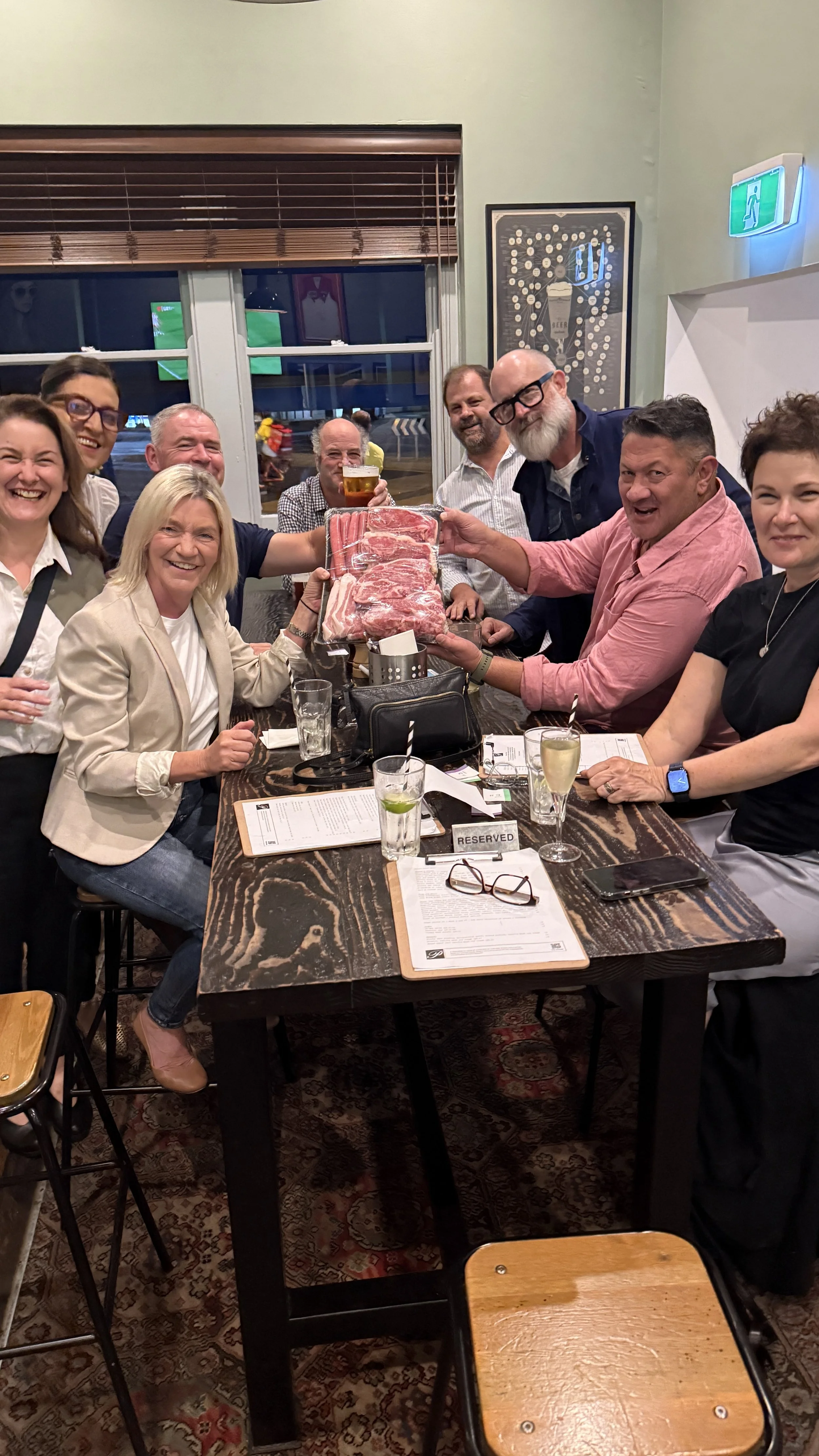Group of people sitting around a restaurant table, holding packages of raw meat, with some holding drinks, smiling and celebrating.