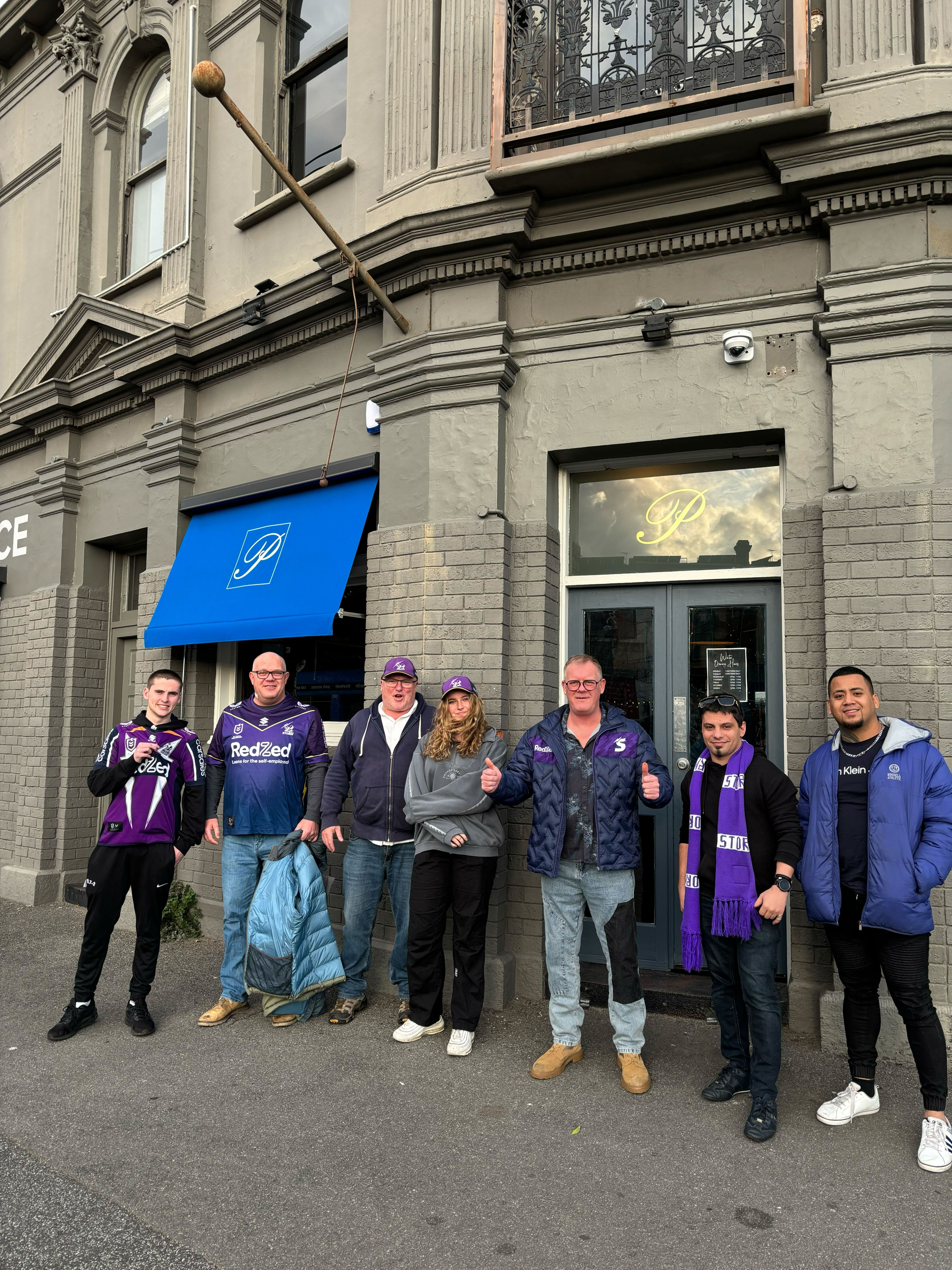 Group of seven people standing outside the Palace Hotel  with a blue awning and a window with a gold logo. The group includes men and women dressed casually, some wearing Melbourne Storm sports jerseys and scarves.