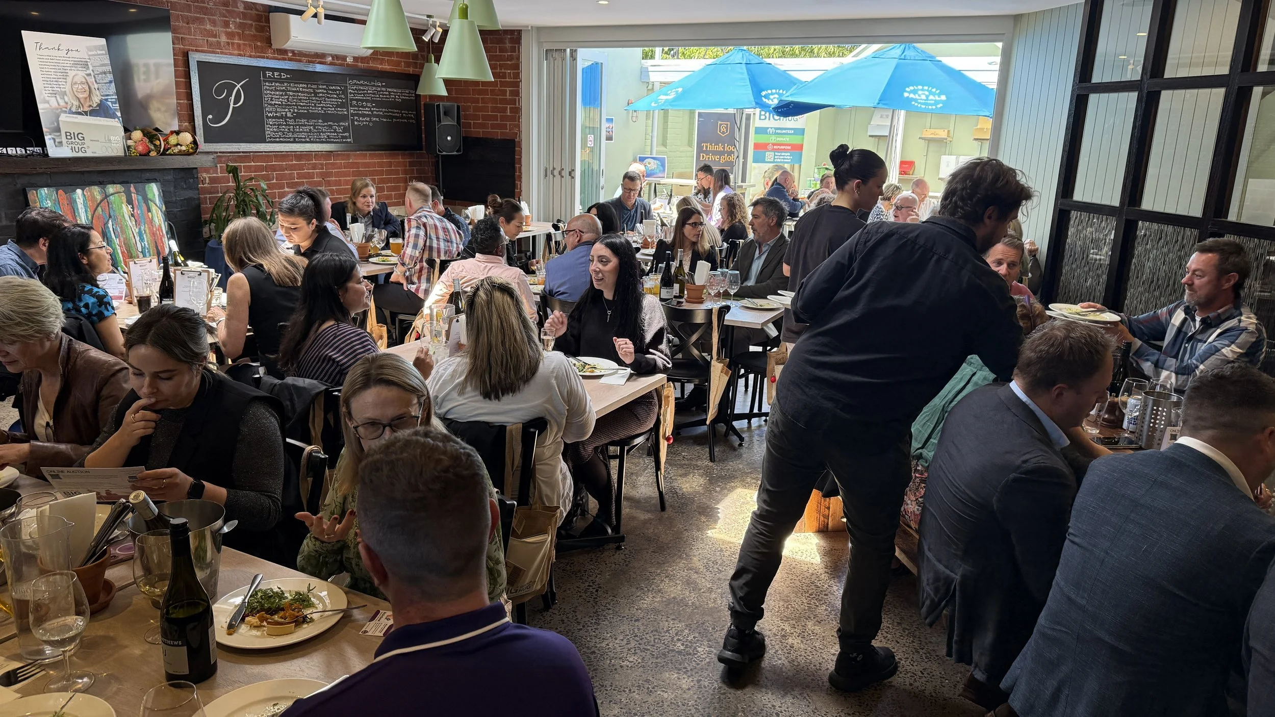 A busy restaurant filled with people enjoying meals and conversation. There are several tables with food, drinks, and glasses, and a server is taking orders. The interior features brick walls, colorful artwork, and large windows with blue umbrellas o