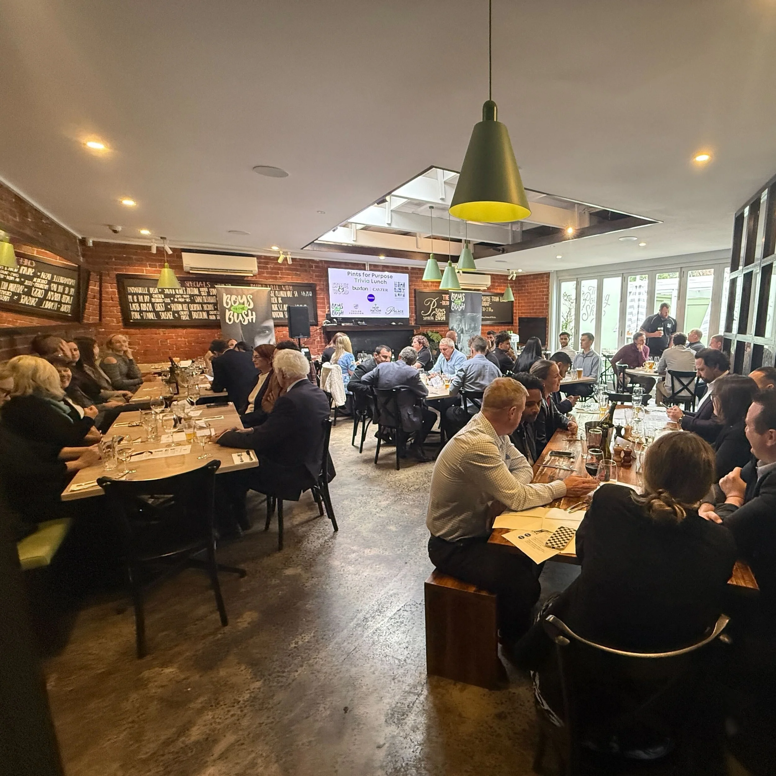 Inside a crowded restaurant with wooden tables and brick walls, people are sitting and dining. There are green pendant lights hanging from the ceiling and a large screen displaying event details.