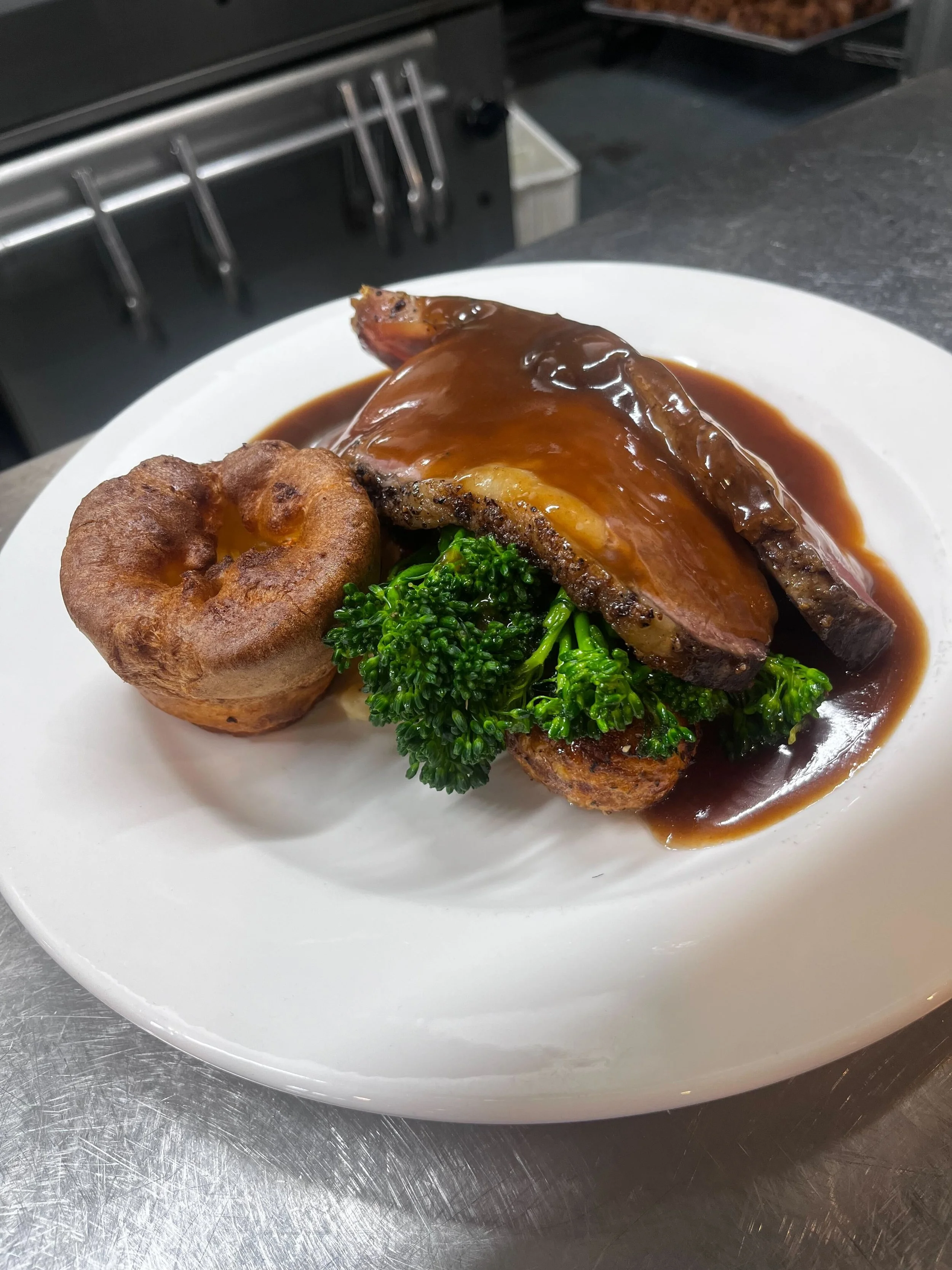 A plate with a cooked ham steak topped with gravy, a small bread roll, and a bunch of broccoli.