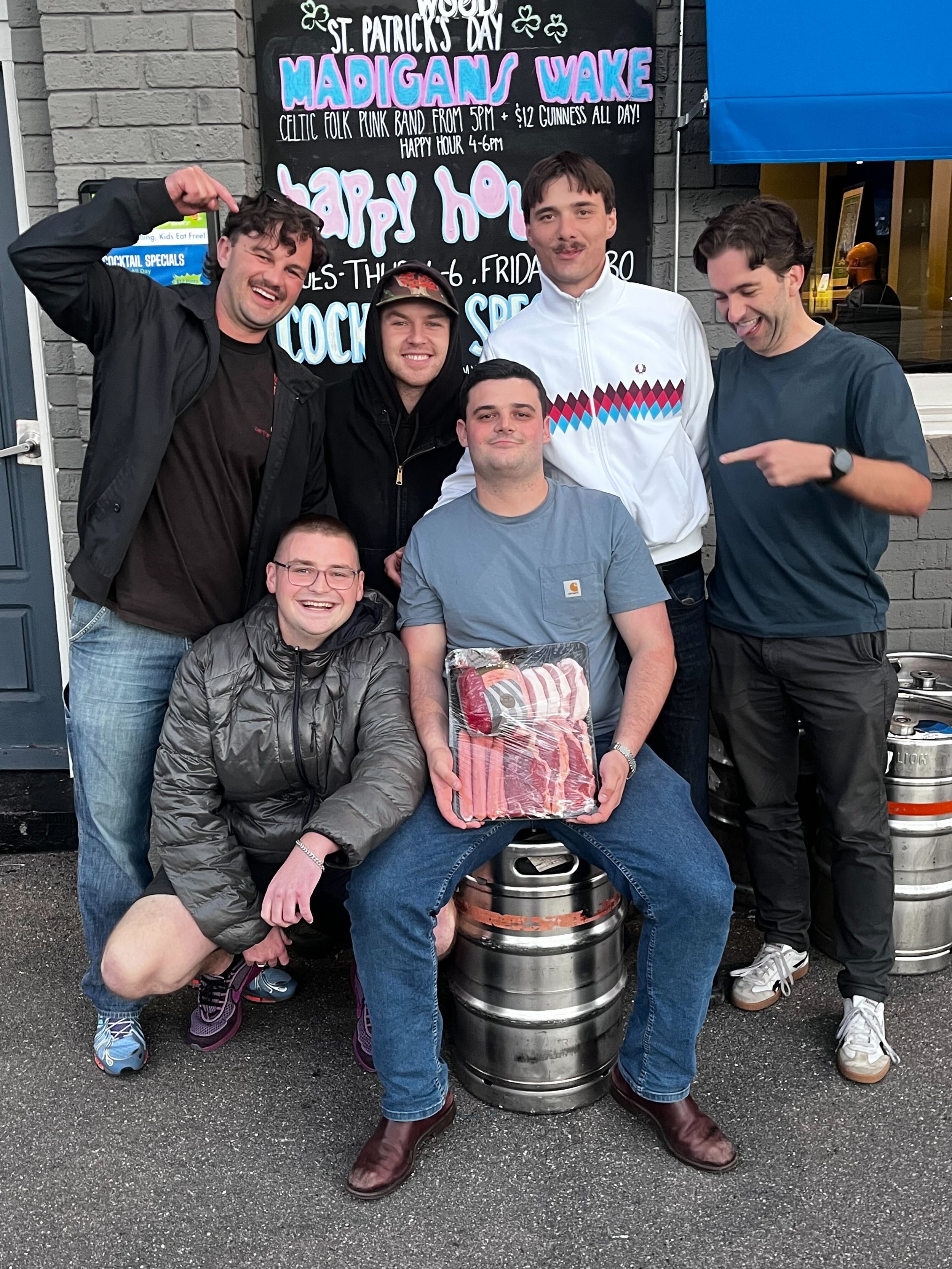 A group of six men smiling and posing in front of a blackboard sign at St. Patrick's Day, with some holding a package of bacon and standing behind beer kegs.