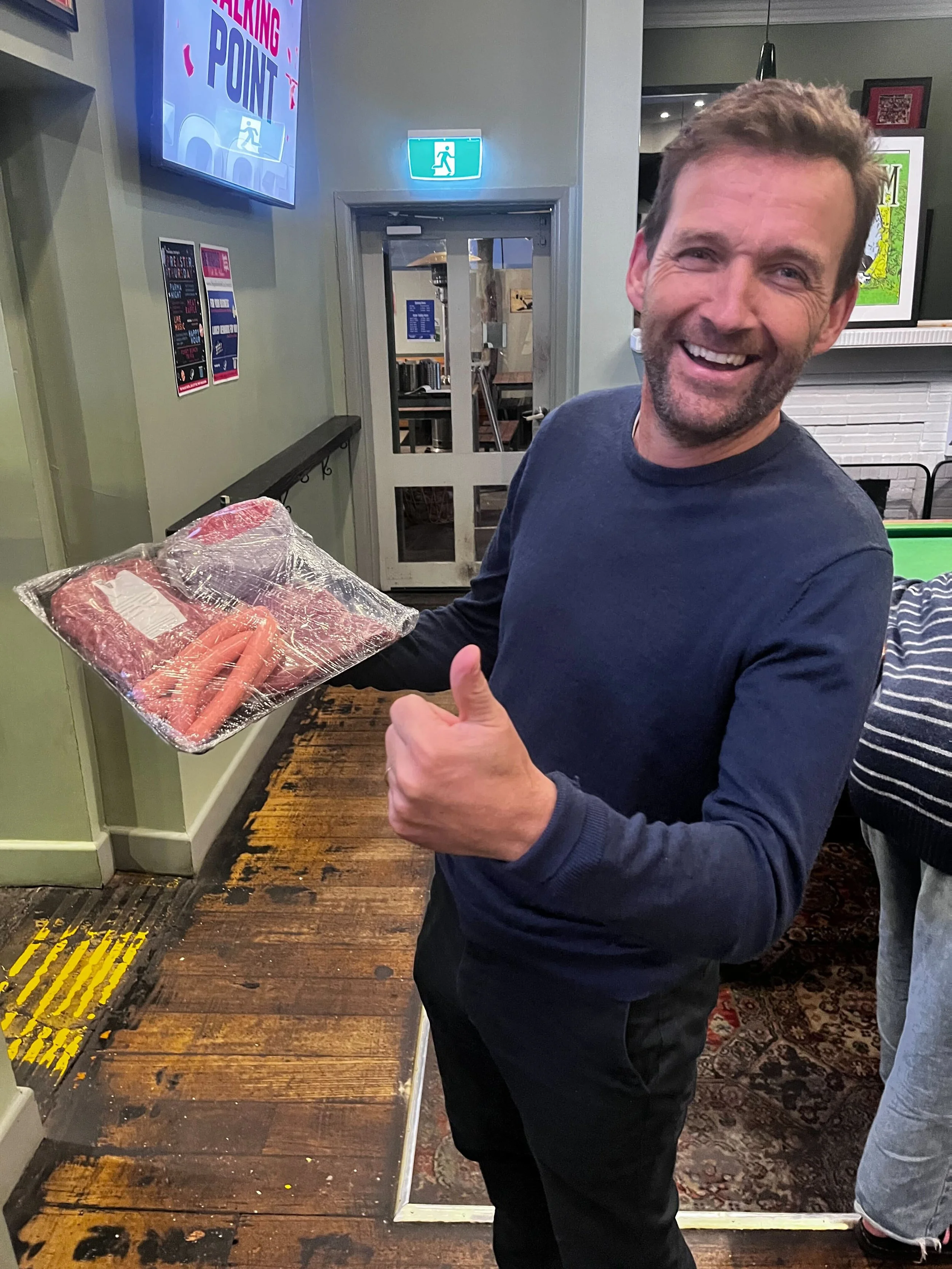 Smiling man holding a package of raw sausages in a restaurant or bar setting, giving a thumbs-up gesture.
