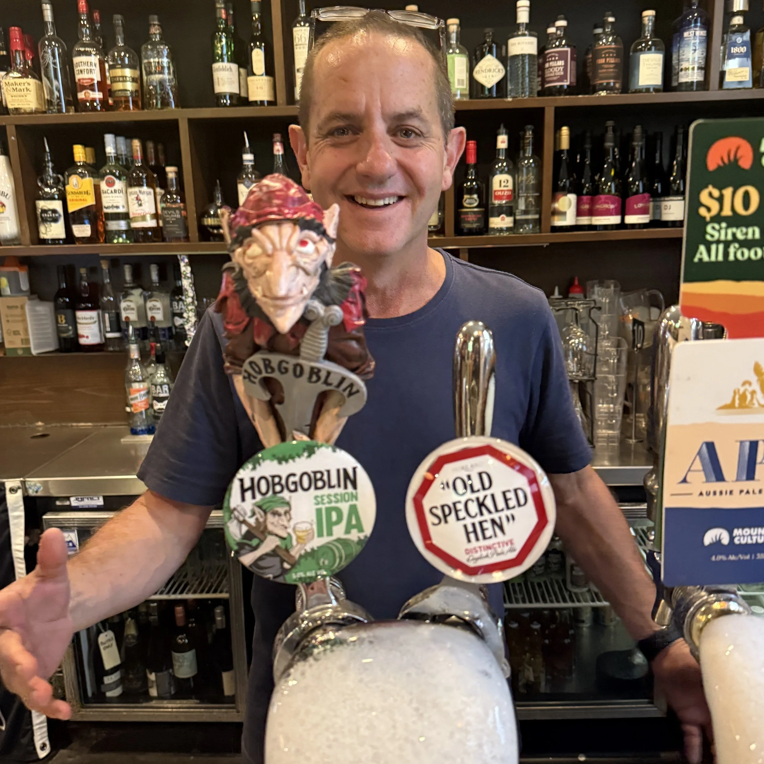 A smiling man behind a bar with two beer taps labeled 'Hobgoblin Session IPA' and 'Old Speckled Hen'.