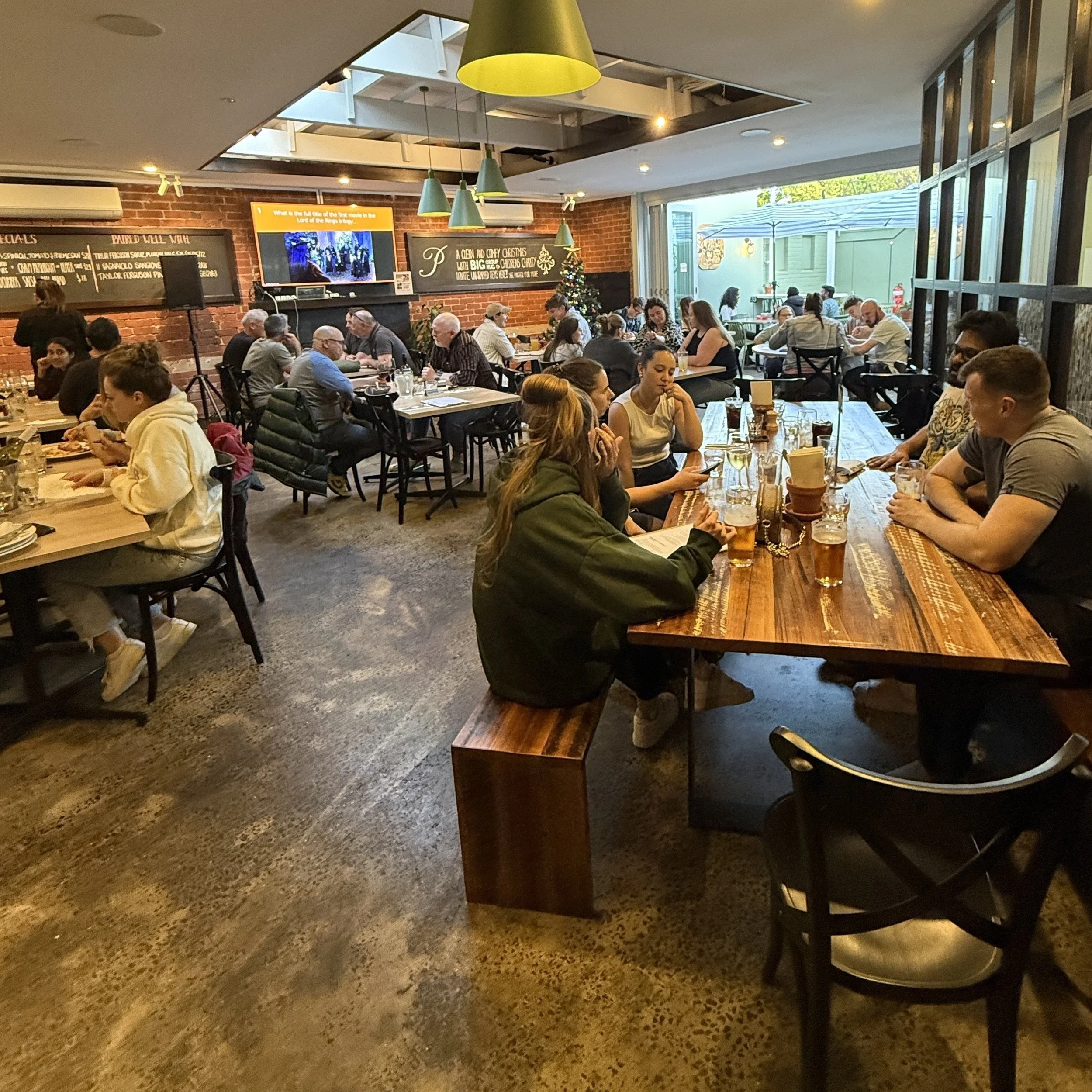 Interior view of a crowded restaurant with wooden tables, black chairs, and a brick wall decorated with a Christmas tree and colorful signs. There are people dining and socializing, with some looking at their phones.