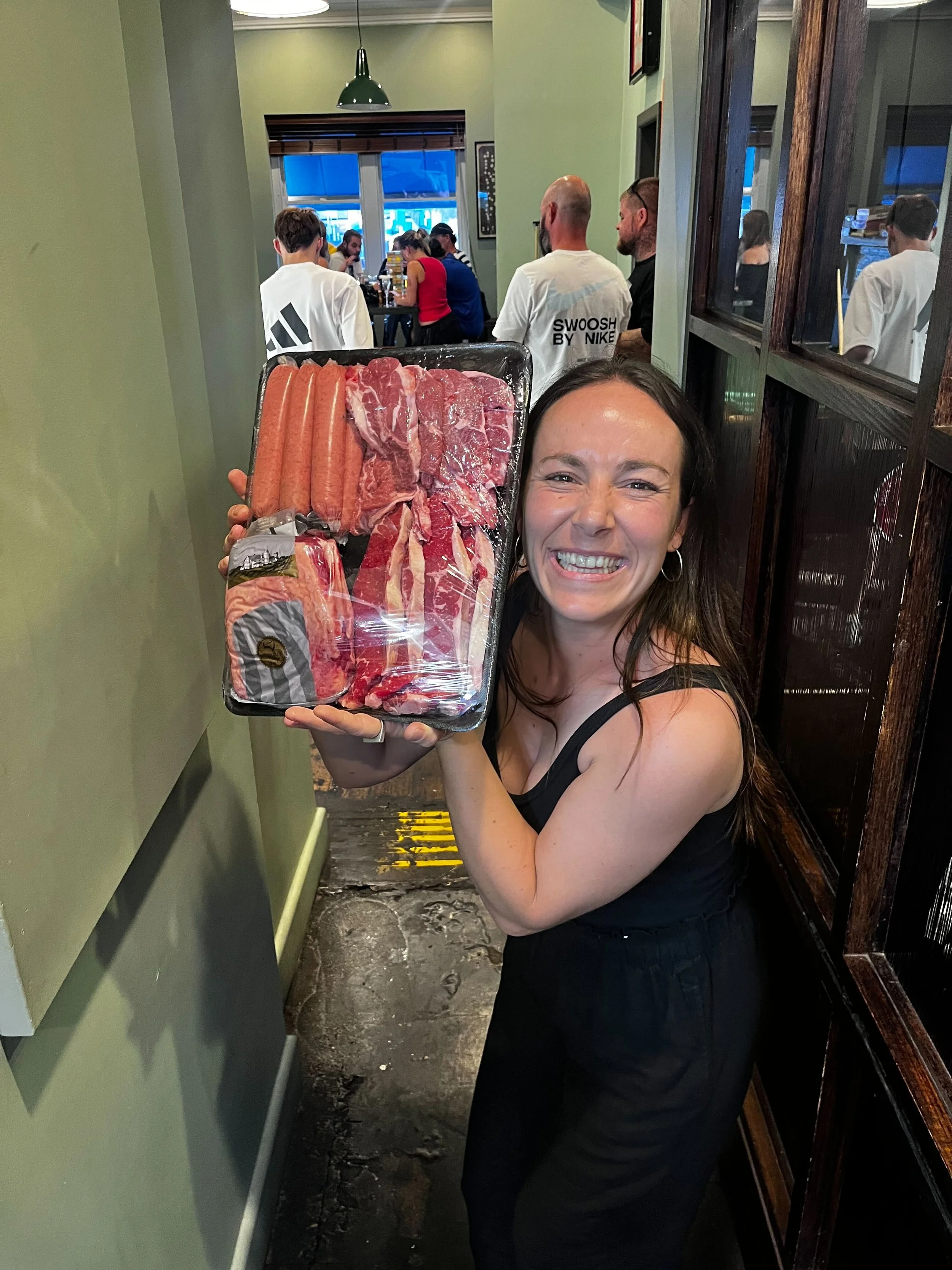 A woman holding a tray of raw meat and sausages inside a restaurant, smiling at the camera as people sit at tables in the background.