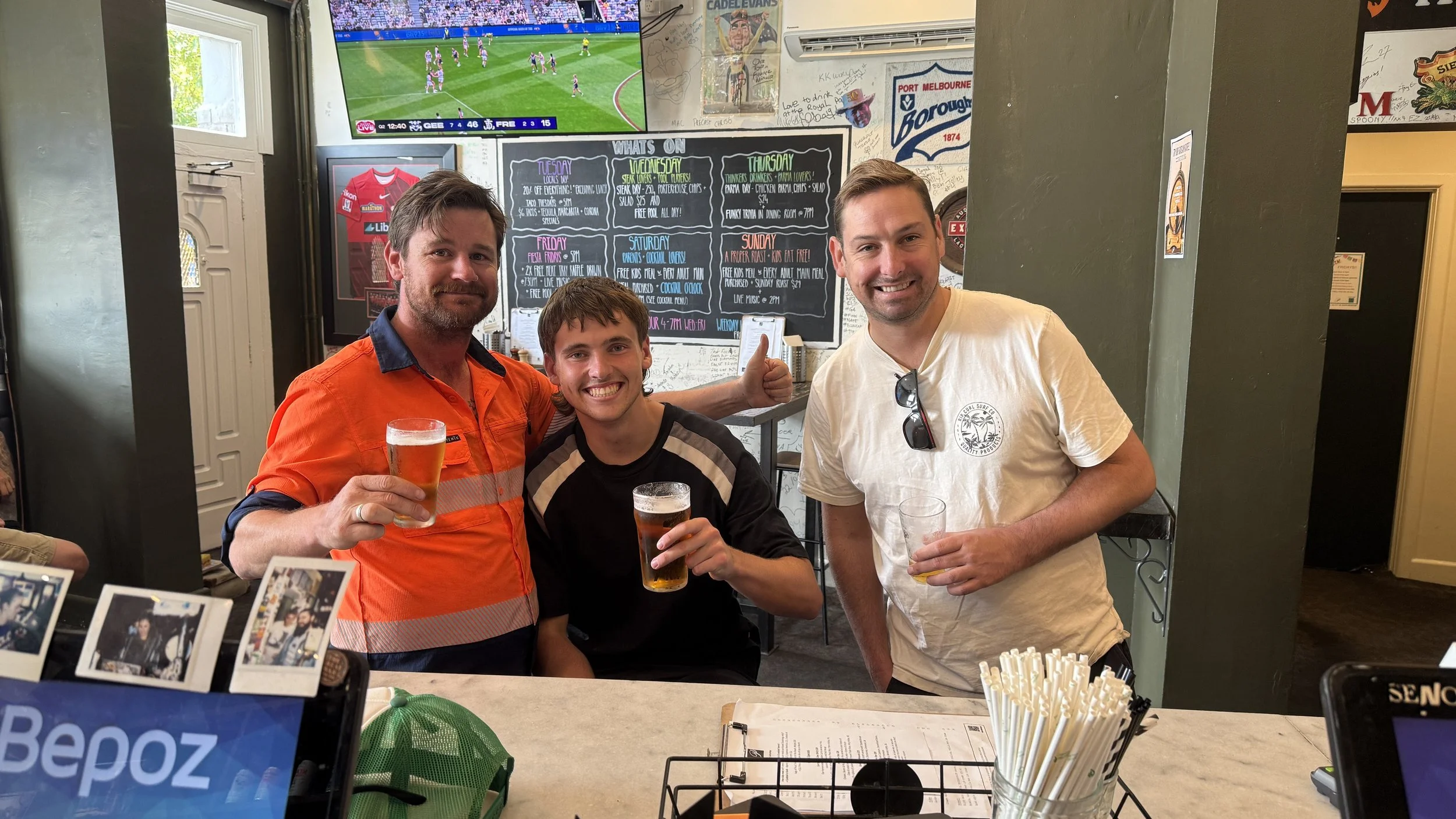 Three smiling men holding up glasses of beer in a bar with a chalkboard menu and television showing a sports game behind them.