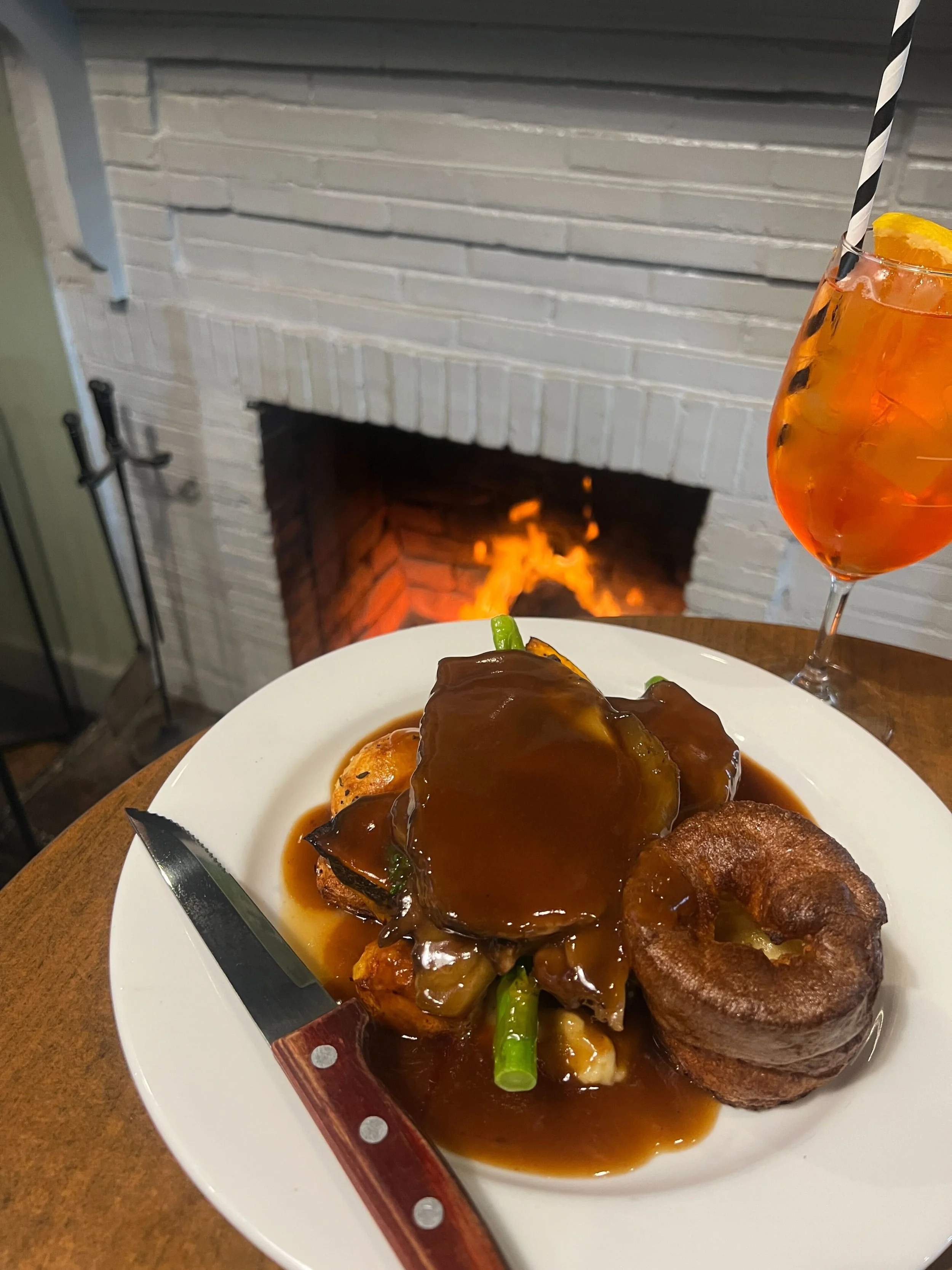Plate of food with a steak topped with brown gravy, roasted potatoes, and green beans, accompanied by an orange cocktail in a tall glass with ice and a lemon slice, set in front of a fireplace.