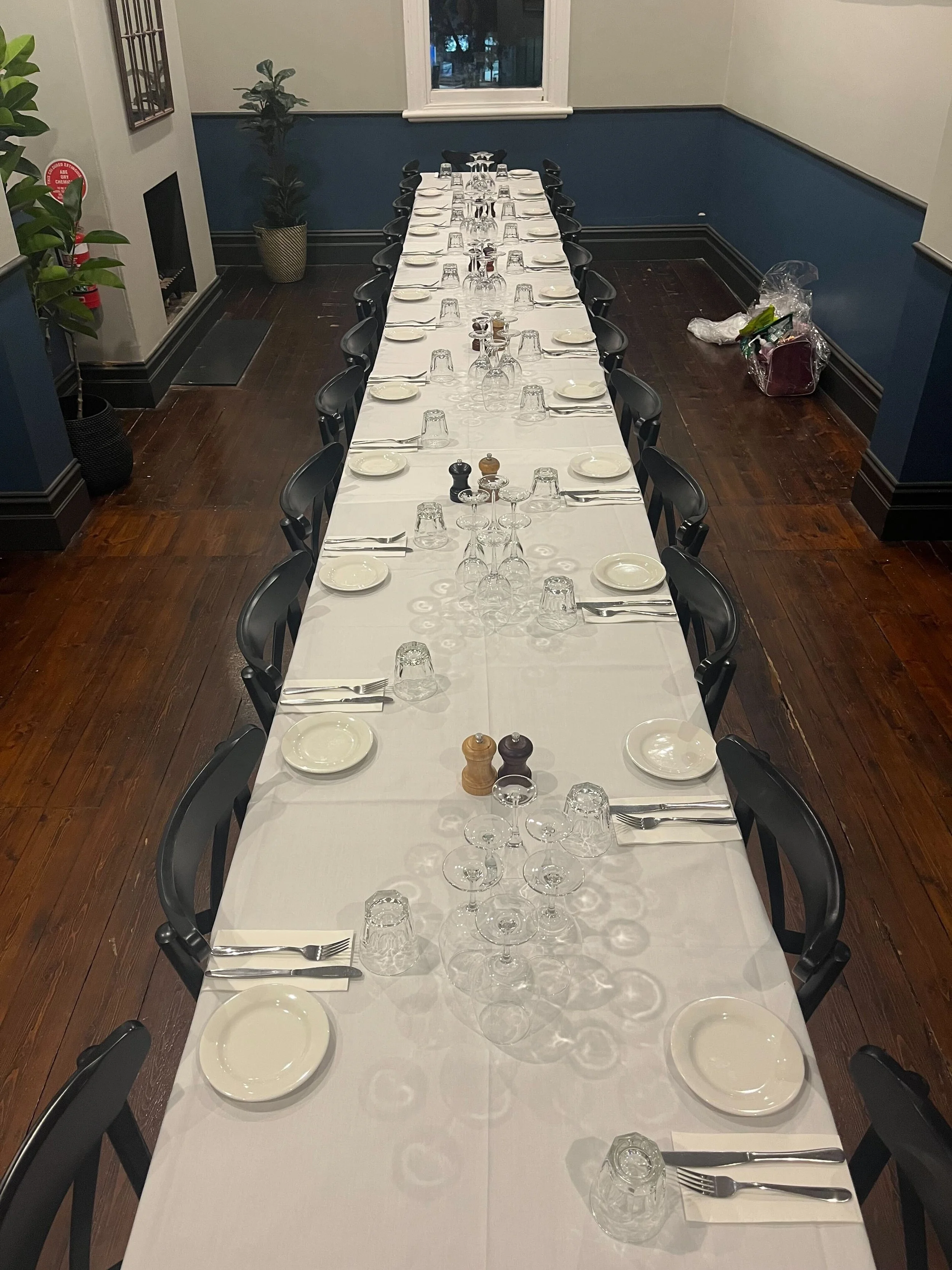 A long dining table set for a formal meal with white tablecloth, black chairs, plates, silverware, upside-down glasses, salt and pepper shakers, and a small centerpiece. The room has dark hardwood floors, a window at the far end, and plants on the si