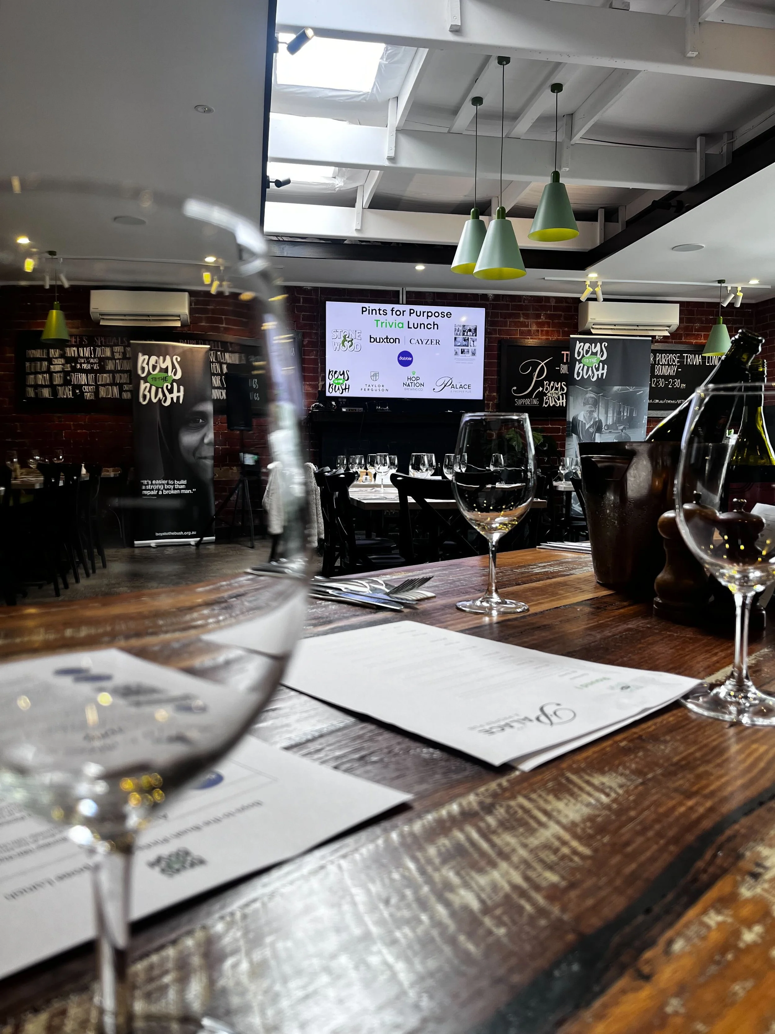 A restaurant setting with tableware, wine glasses, and menus on a wooden table amid a dinner event called 'Pints for Purpose.' Decor includes hanging green pendant lights and promotional banners for 'Boys to the Bush.'