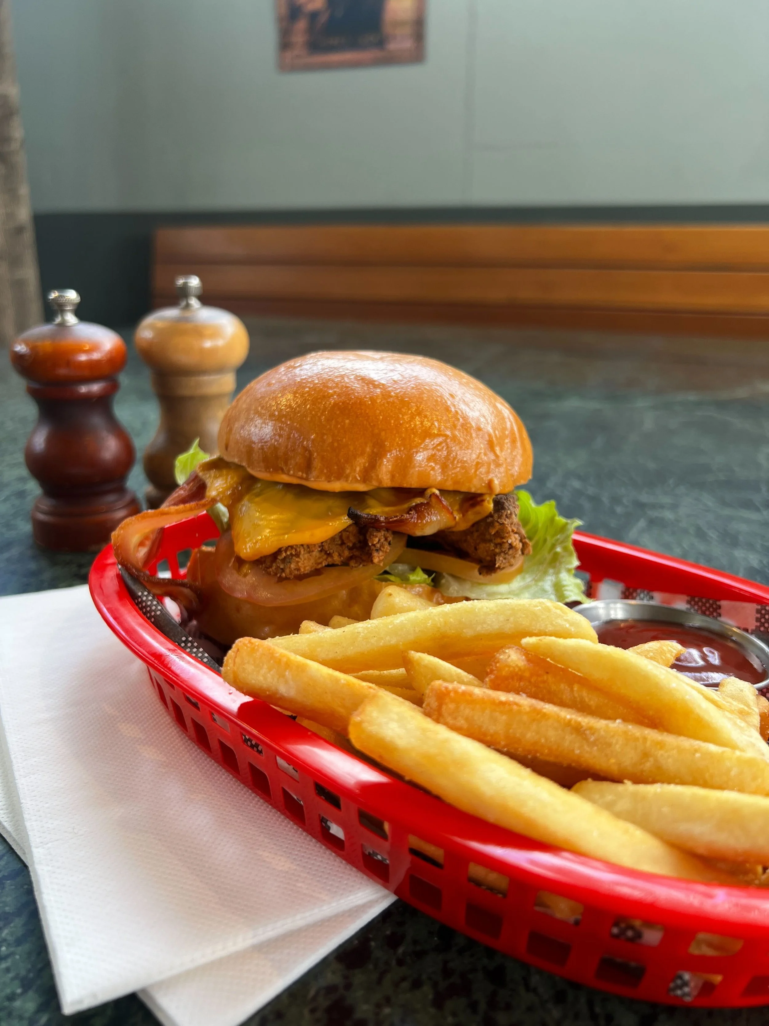 Close-up of a cheeseburger with bacon, lettuce, tomato, and melted cheese in a bun, served with French fries and ketchup in a red basket on a table at a restaurant.