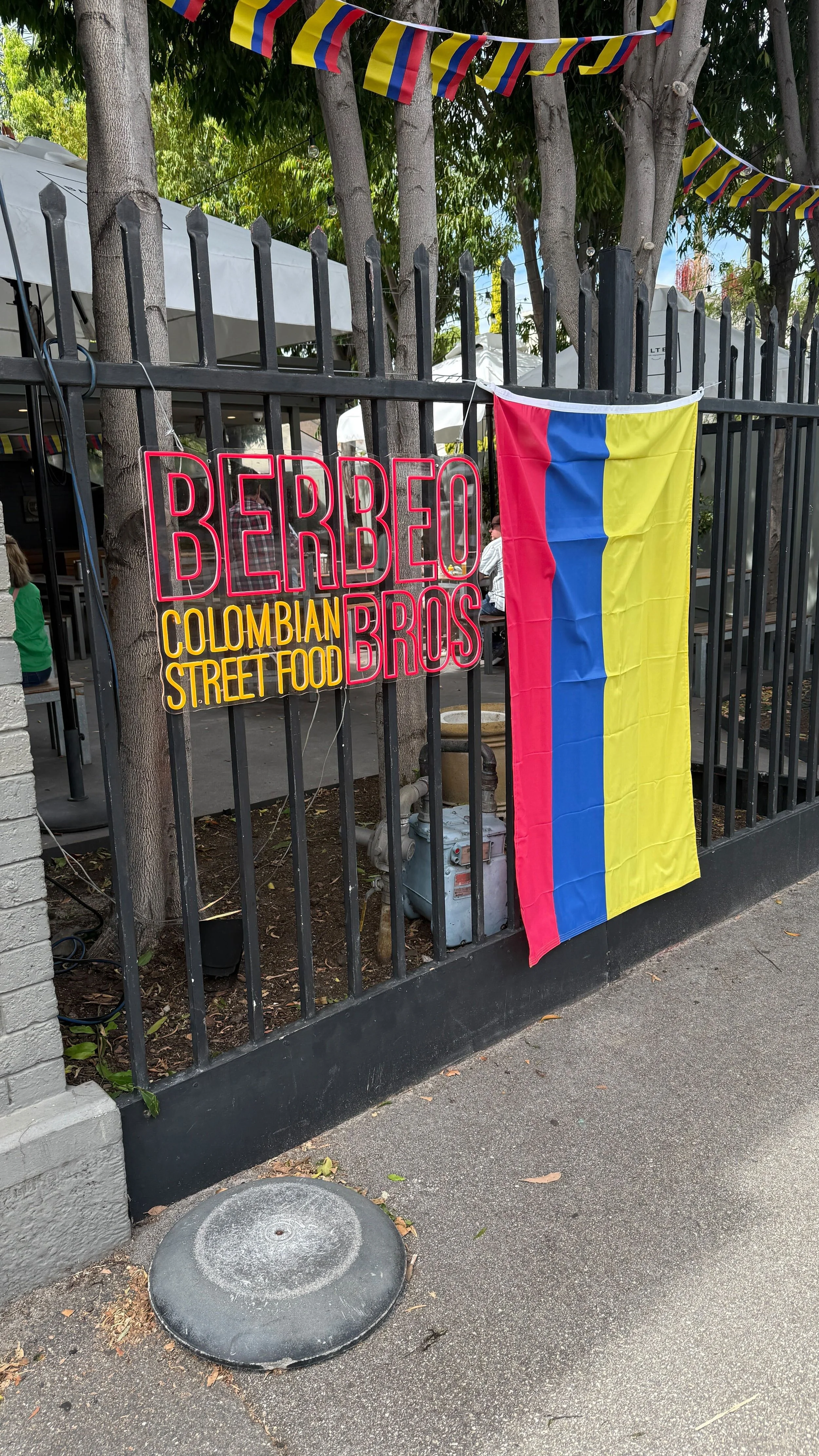 A street food restaurant sign for 'Berbego Bros' at a Colombian street food establishment. A Colombian flag is hung on the black metal fence, with colorful pennant banners above the scene. The sidewalk and part of the restaurant’s outdoor seating are