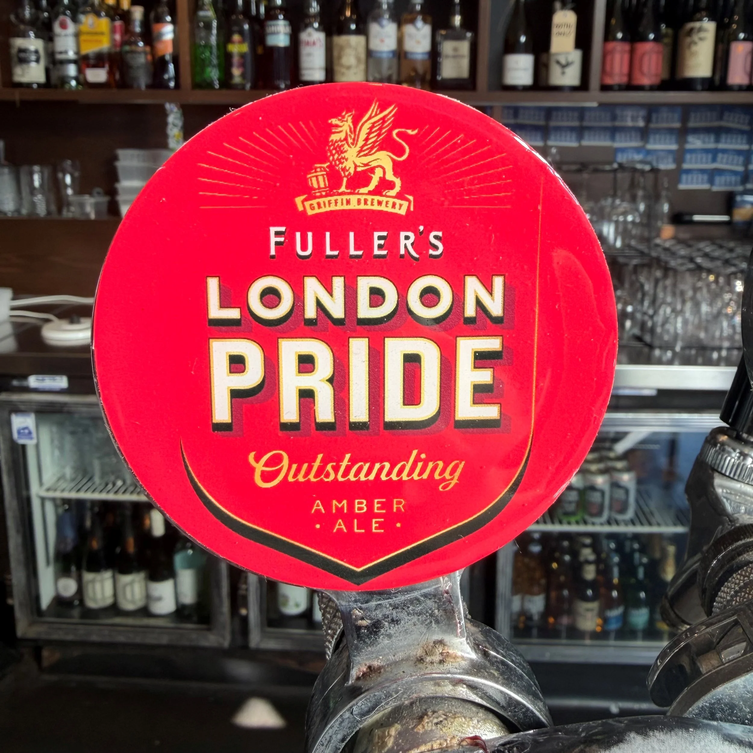 A round red sign from Griffin Brewery advertising London's Pride Amber Ale, featuring a gold griffin logo and the words 'Outstanding' at the bottom, displayed in front of shelves with liquor bottles.