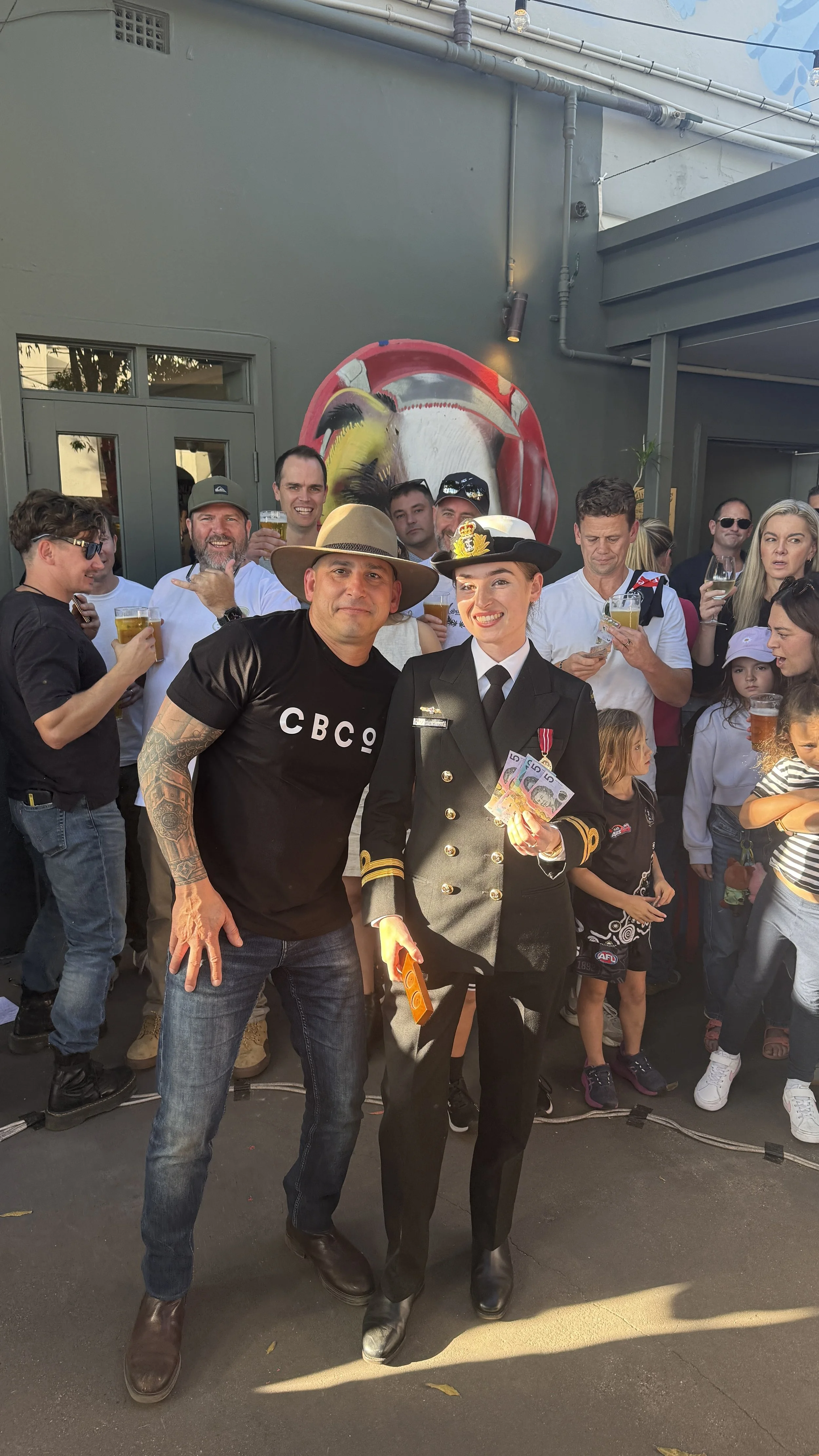 A group of people at a celebration, with two individuals in the foreground: a man wearing a black CBCo shirt and a woman dressed as a police officer holding Australian currency. Other people in the background are holding drinks, and there is a mural 