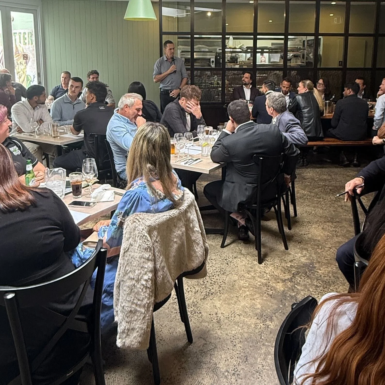 A group of people gathered in a restaurant or cafe, sitting at tables with drinks and eating utensils, listening to a man standing with a microphone, speaking to the audience. The room has large windows and an interior with a mix of modern and rustic decor.