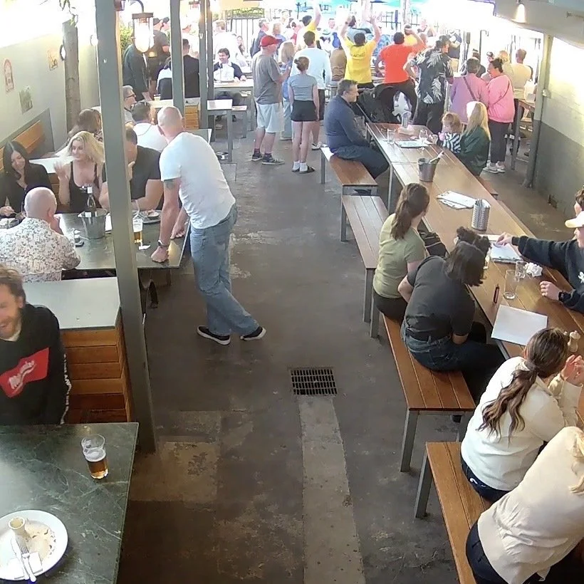 A busy indoor bar or restaurant with many people sitting and standing, enjoying drinks and food. Patrons are socializing at tables and at the bar, with some waiting in line or ordering. The atmosphere is lively and casual.