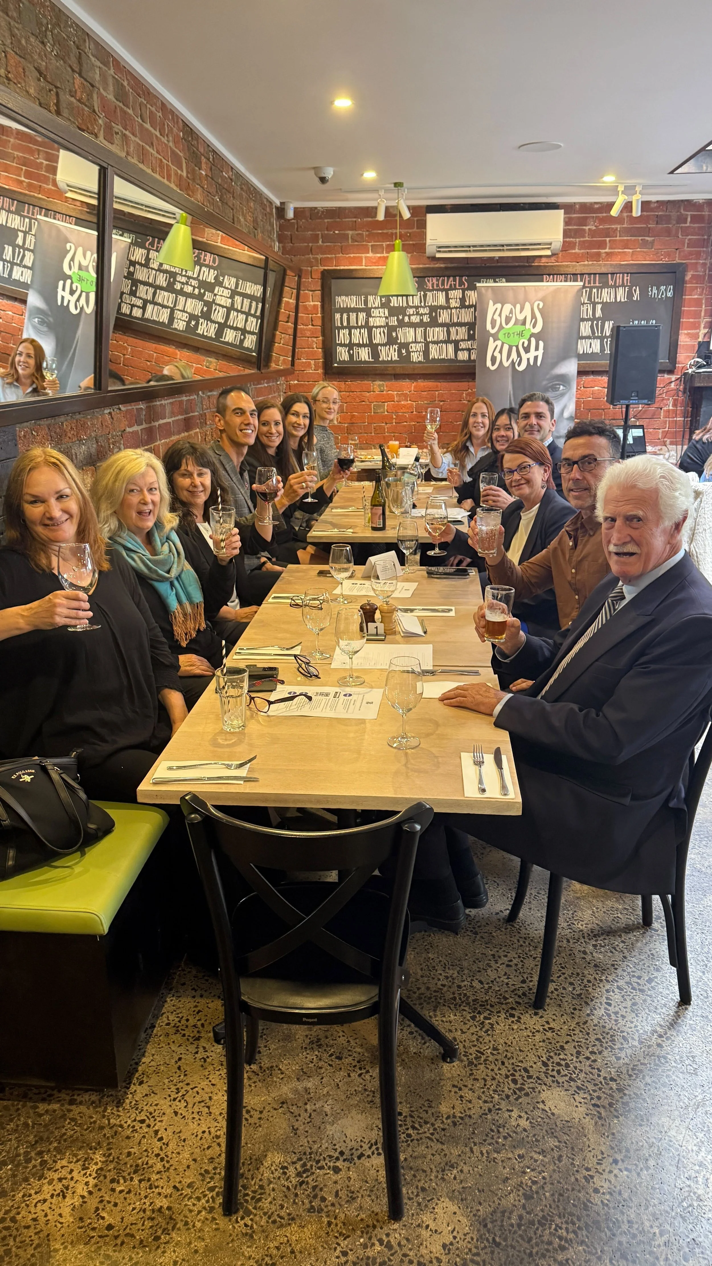 A group of people sitting around a long dining table in a restaurant, smiling and holding glasses of drinks, with exposed brick walls and holiday posters in the background.