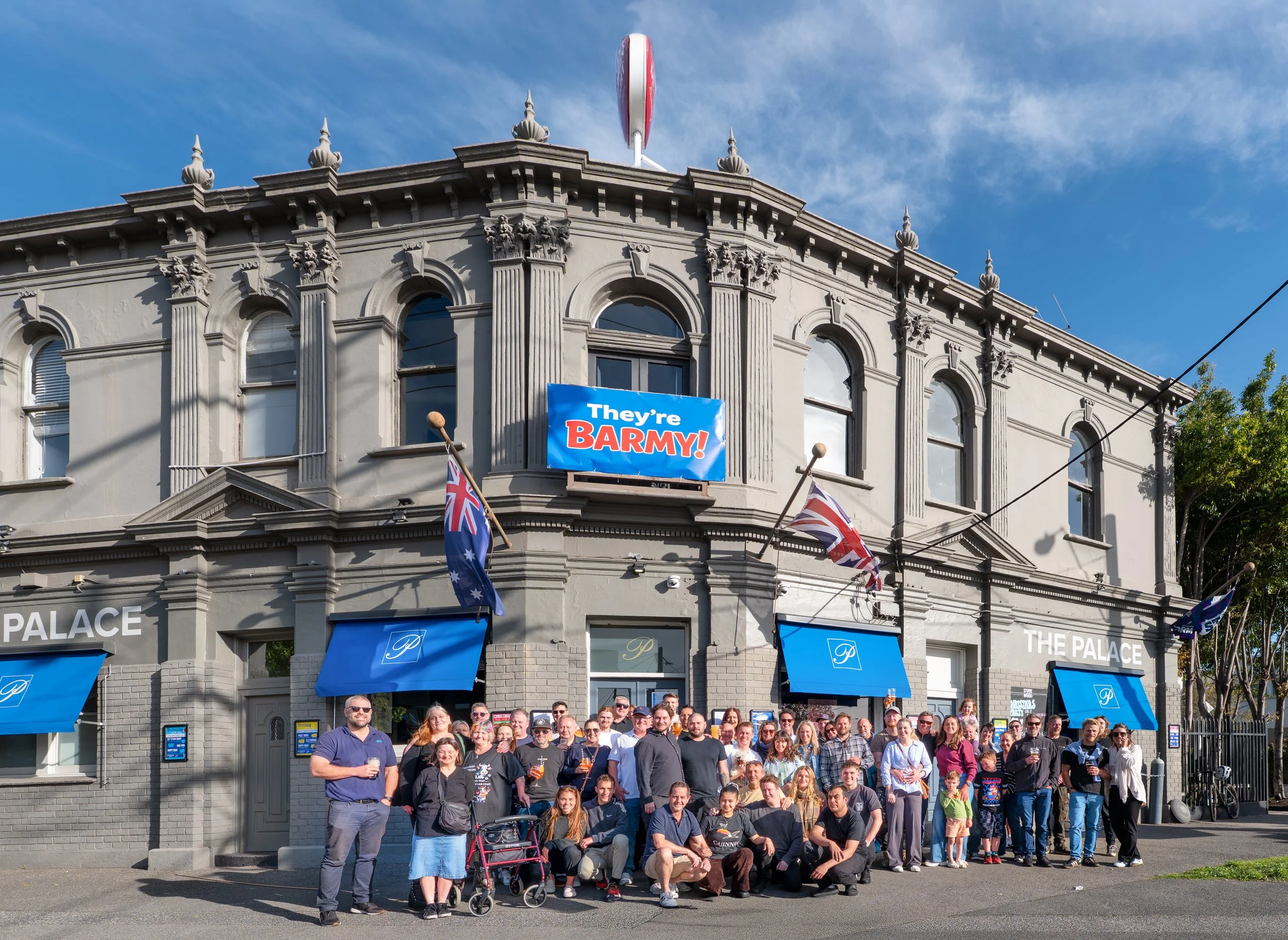 A large group of people gathered in front of a historic building with ornate architectural details, blue umbrellas, and flags, celebrating or attending an event.