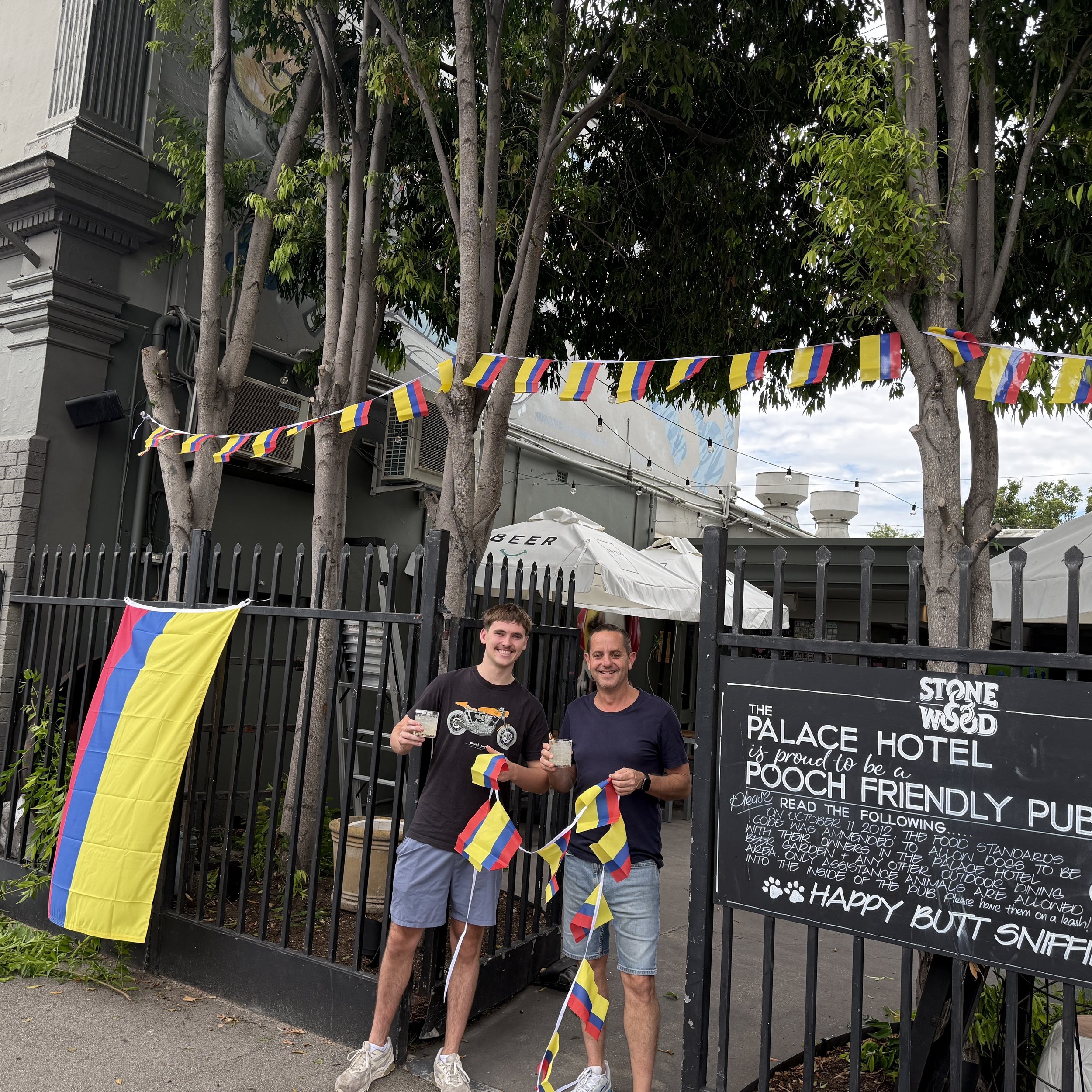 Two men standing outside behind a black fence, smiling and holding drinks, with flags and a blackboard sign nearby, and trees overhead.