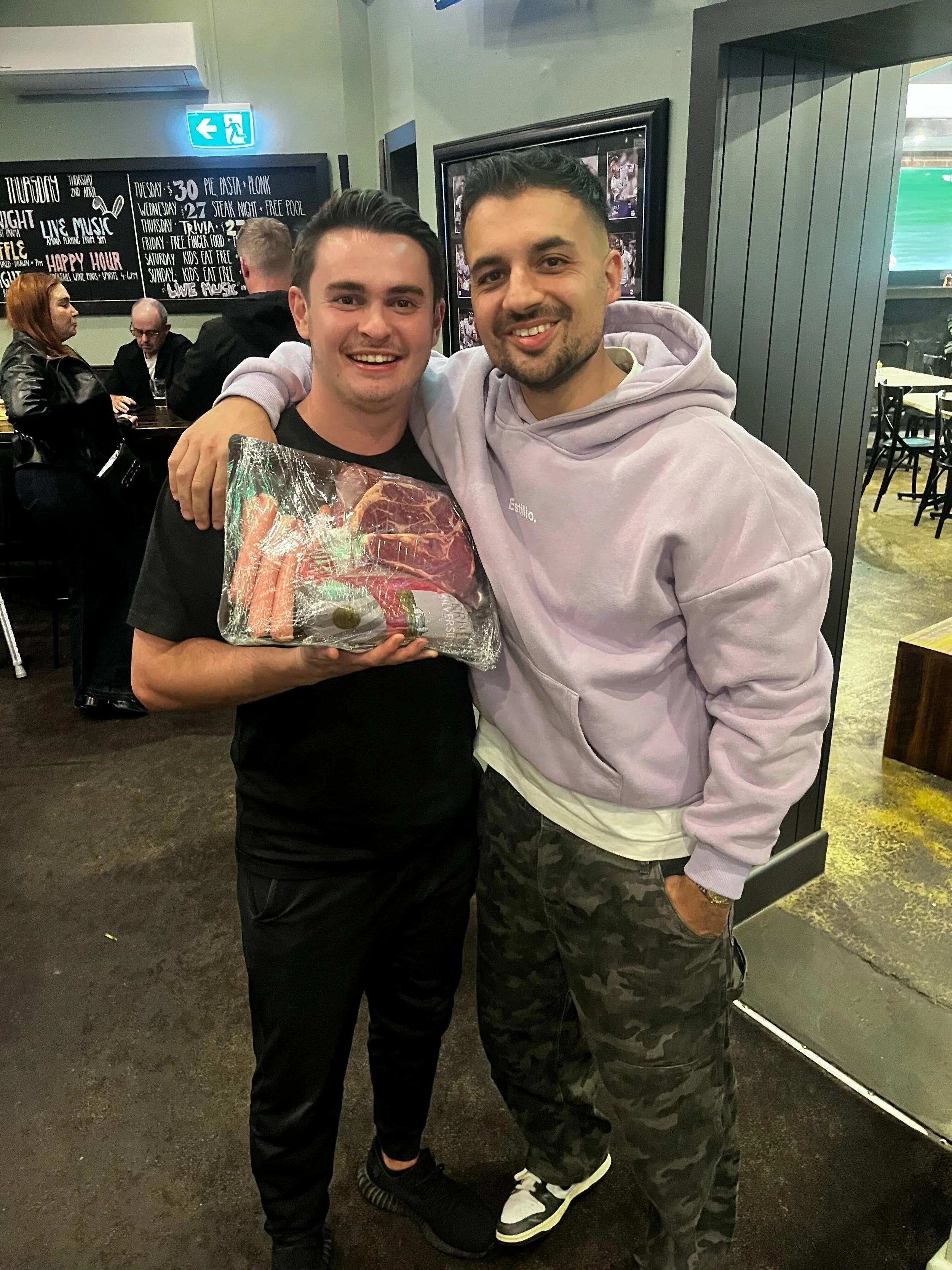 Two young men are smiling and posing together in a casual restaurant or bar. One is holding a wrapped gift. The background features a blackboard menu and other patrons seated at tables.