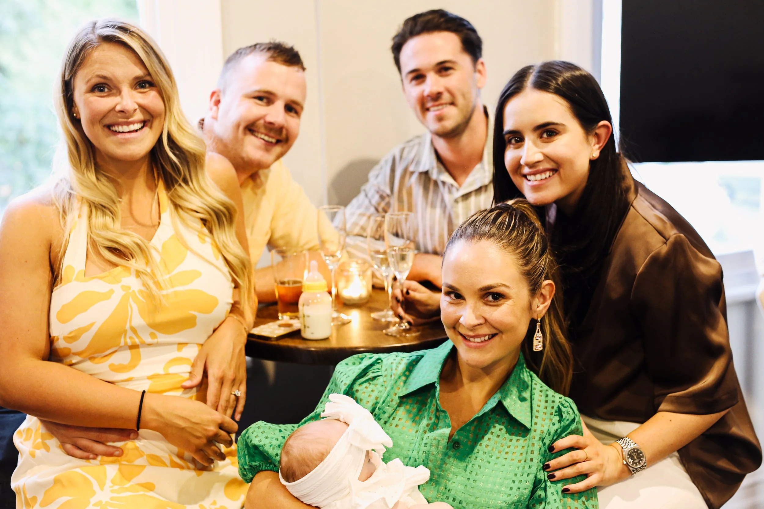 Group of six people, including a woman holding a newborn, gathered around a table at a celebration in a cozy indoor setting.