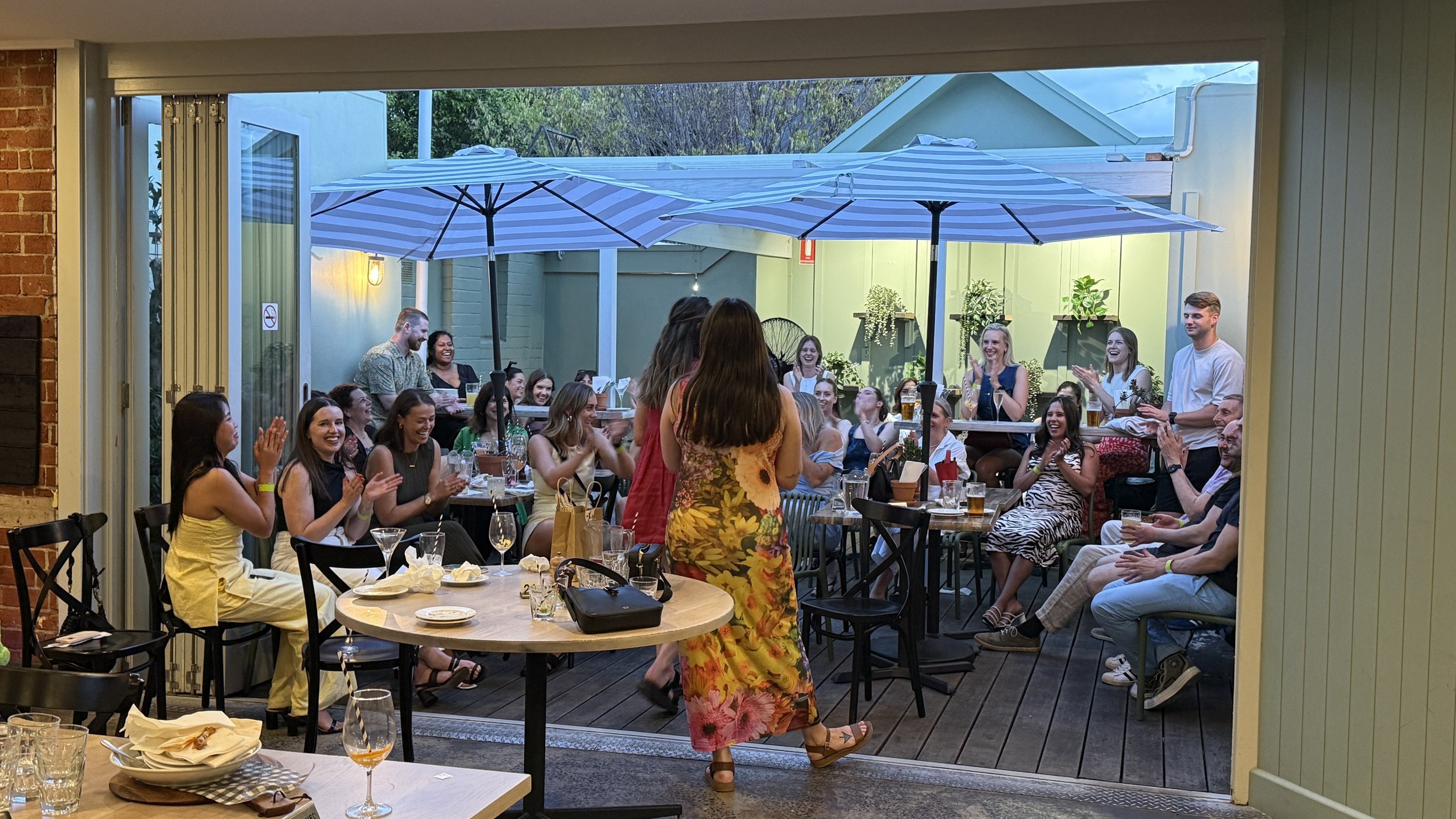 People gathered at a party on a patio with large umbrellas, enjoying drinks and laughing.