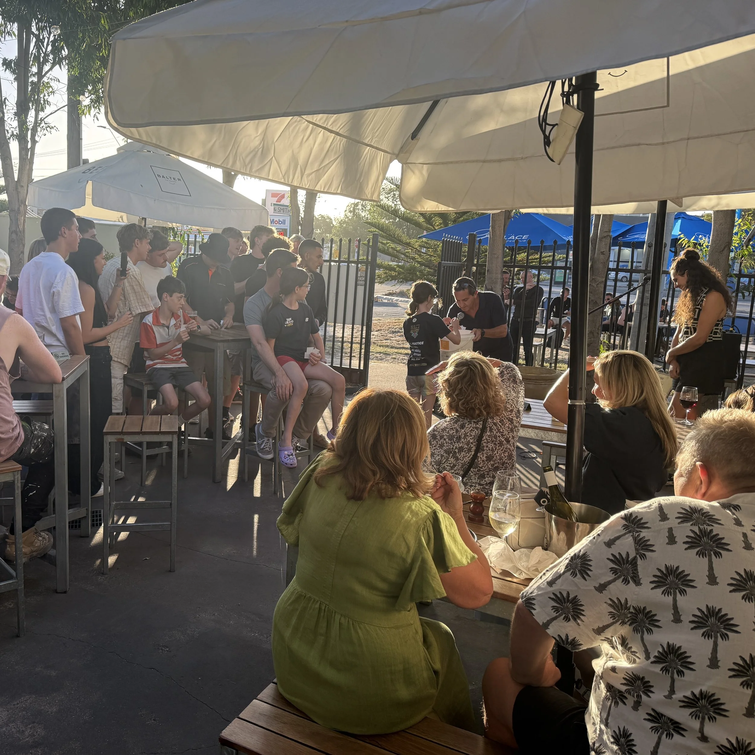 People gathered outdoors under umbrellas, sitting and standing around tables, at a social event during late afternoon or early evening.