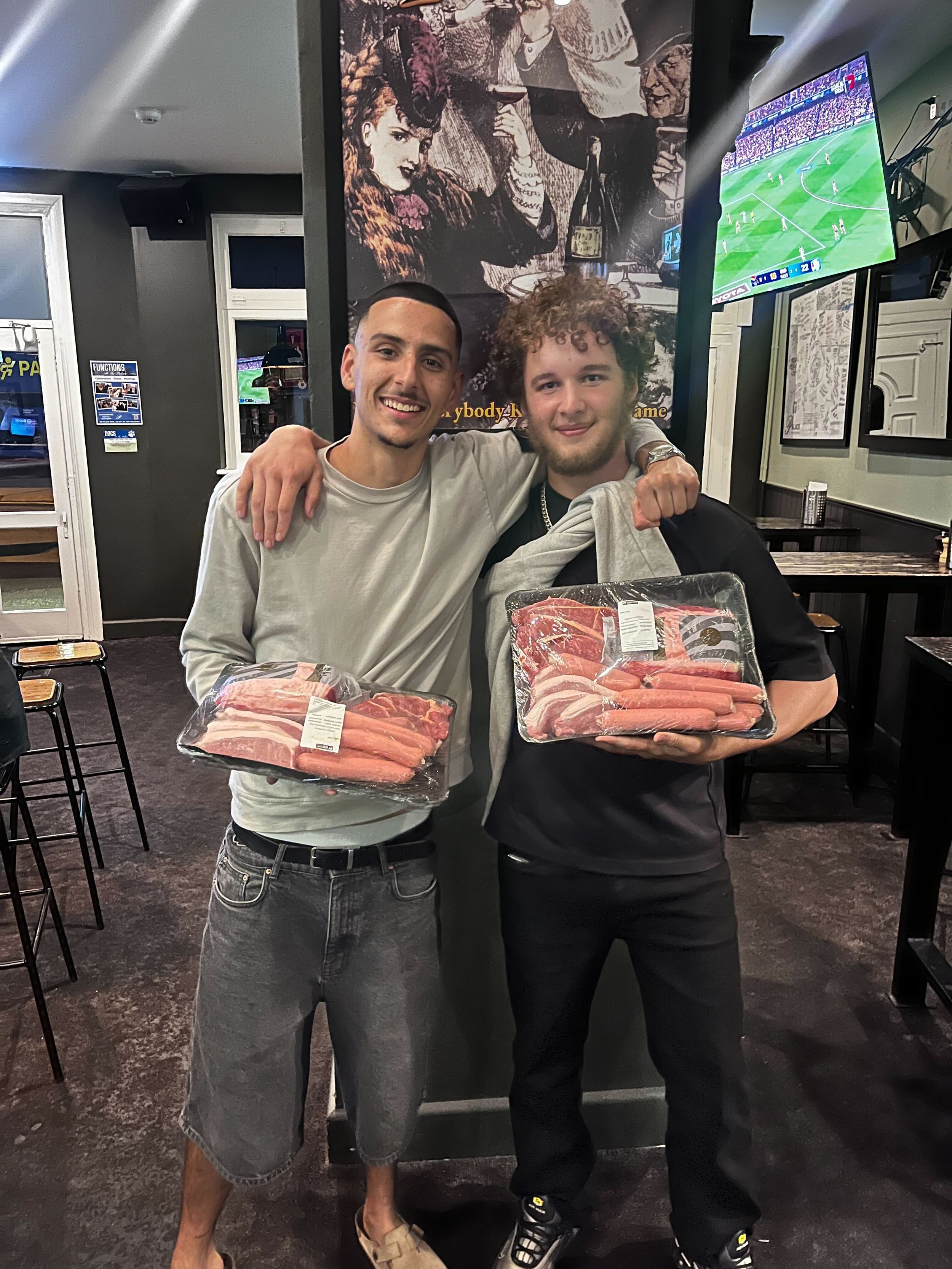 Two young men standing in a restaurant holding packages of raw meat, with arms around each other and smiling at the camera.