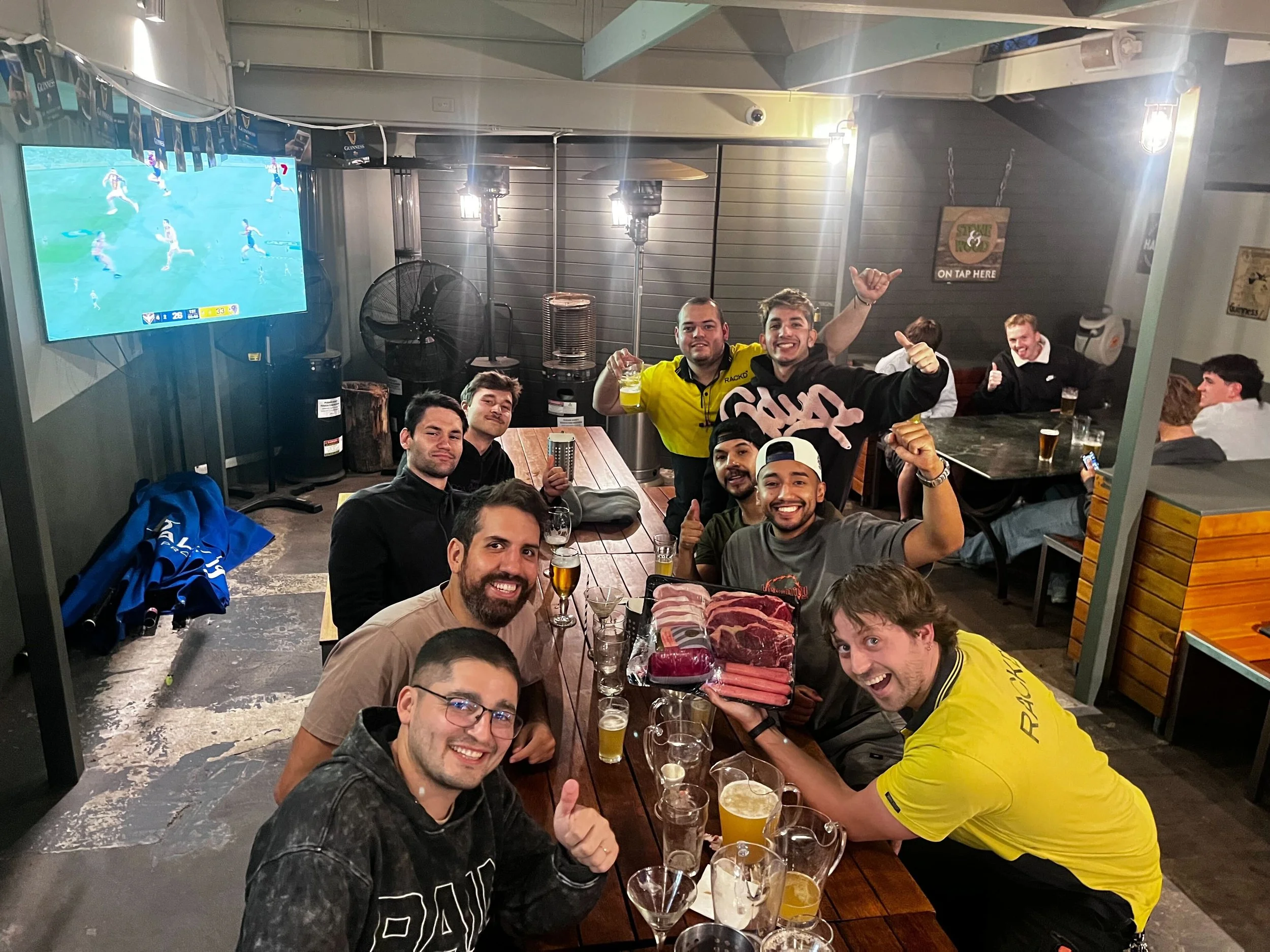 A group of smiling men sitting and standing around a wooden table in a bar or restaurant, holding drinks and giving thumbs-up signs. There are various beverages, including beer and a beer mug with a foam head, on the table. In the background, a flat-