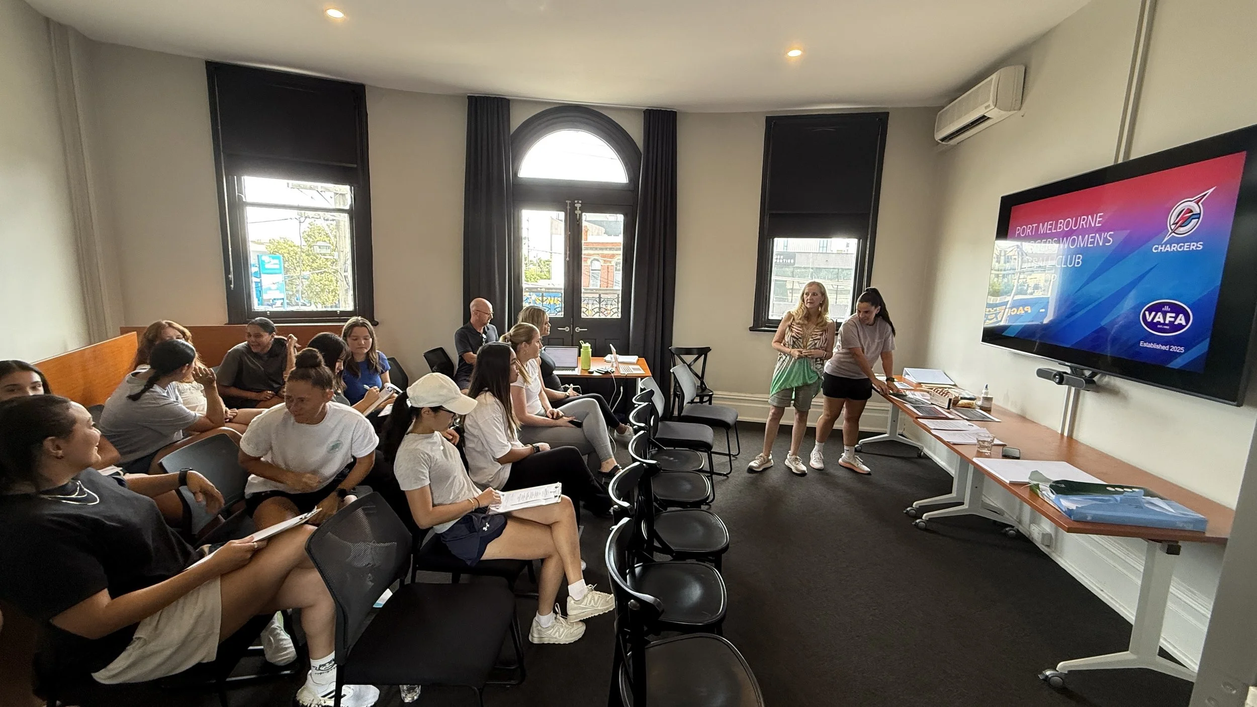 Group of young women and girls seated in a classroom or meeting room, listening to a presentation about women's basketball. There is a large screen with the logo of the Port Melbourne Chargers Women's Football Club.