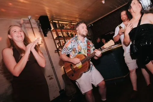 A group of people, including a woman clapping and a man playing guitar, enjoying music at a social gathering or bar. There are two other individuals in the background, one with a drink, in a dimly lit indoor environment.