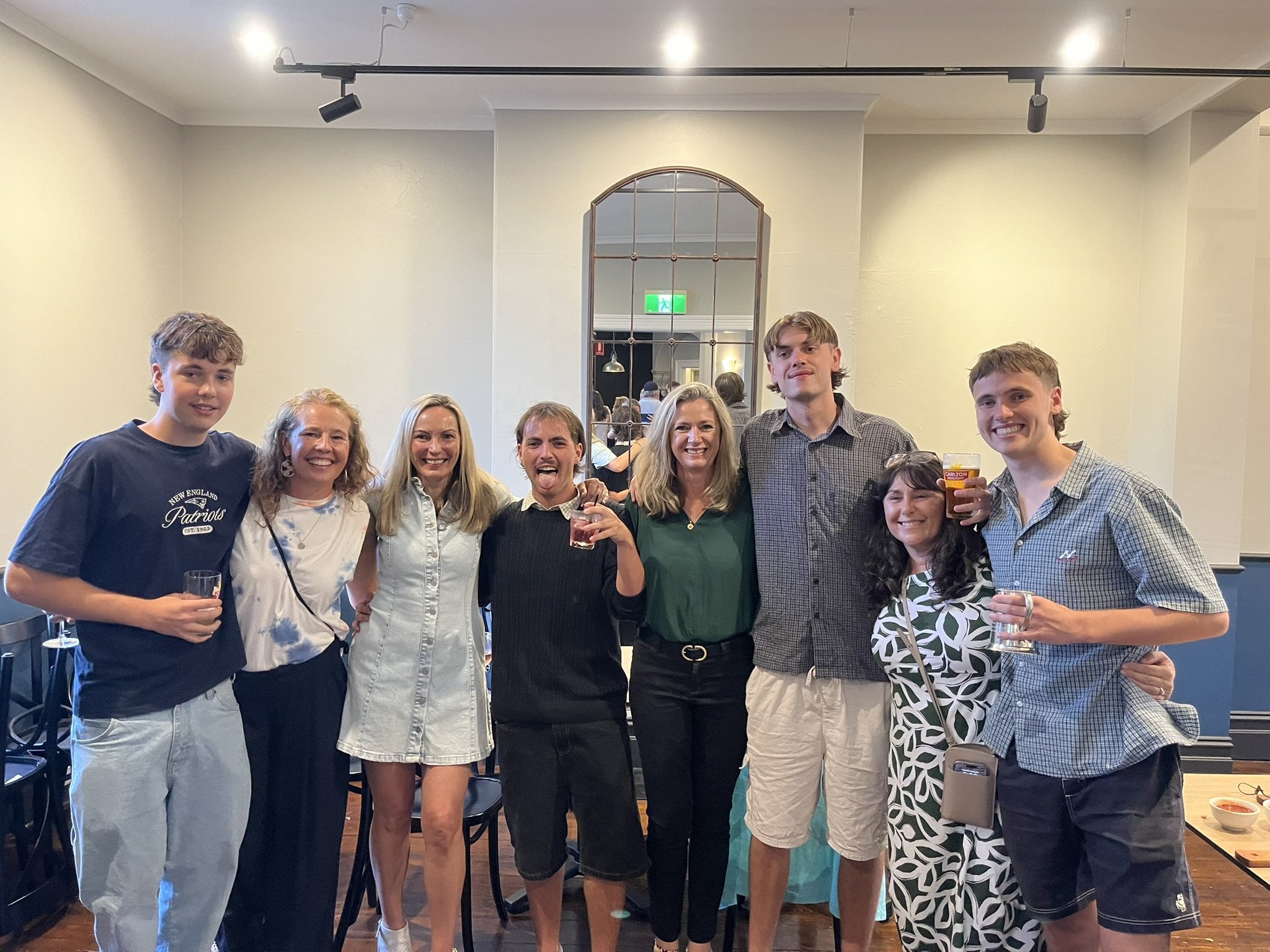 A group of nine people smiling and posing together at a birdthday party upstairs at the Palace Hotel , some holding drinks, indoors with a mirror and an exit sign in the background.