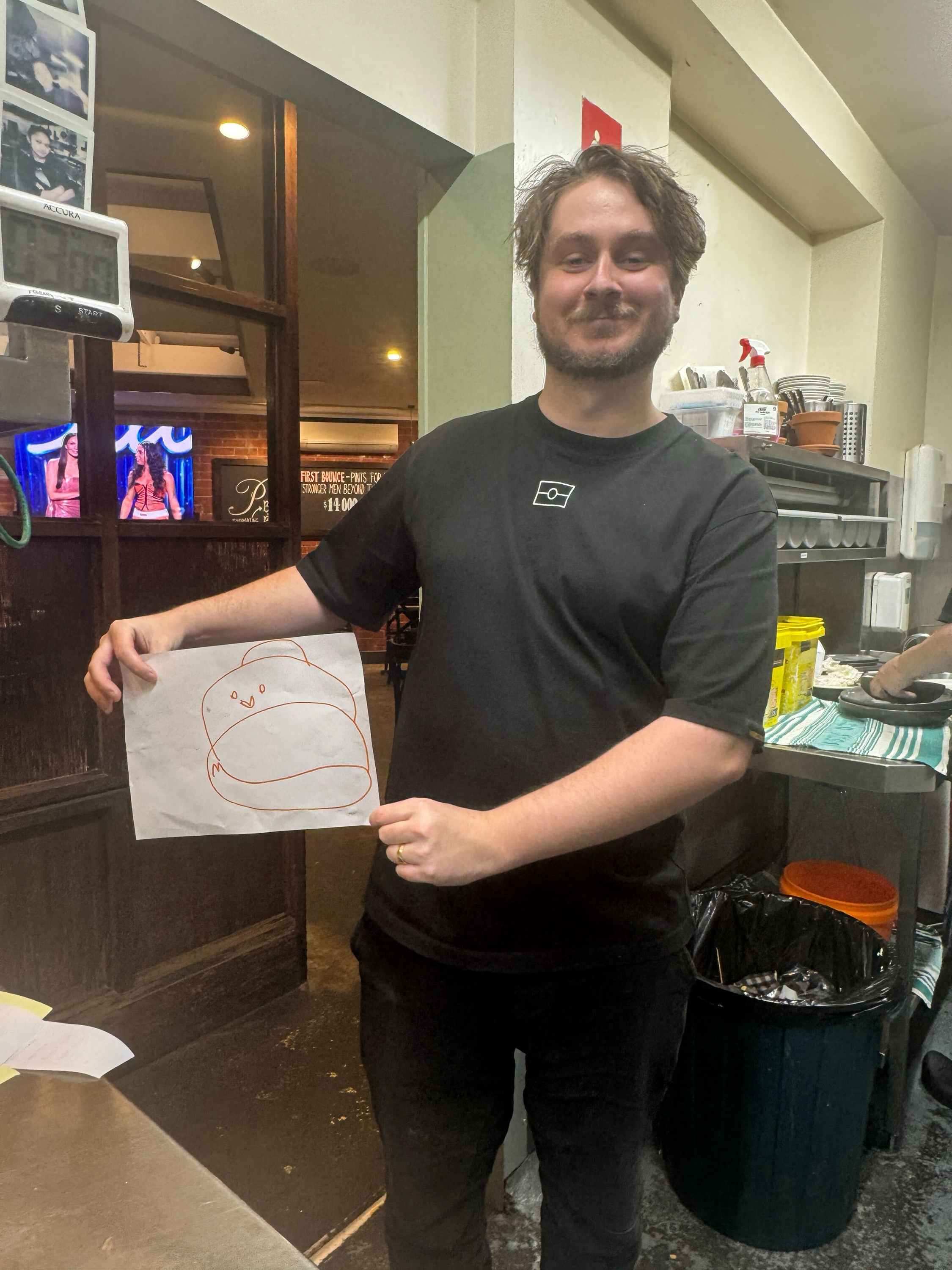 A person with curly hair and a beard, smiling and holding a drawing of a chick, standing in a restaurant or cafe with kitchen equipment and tables in the background.