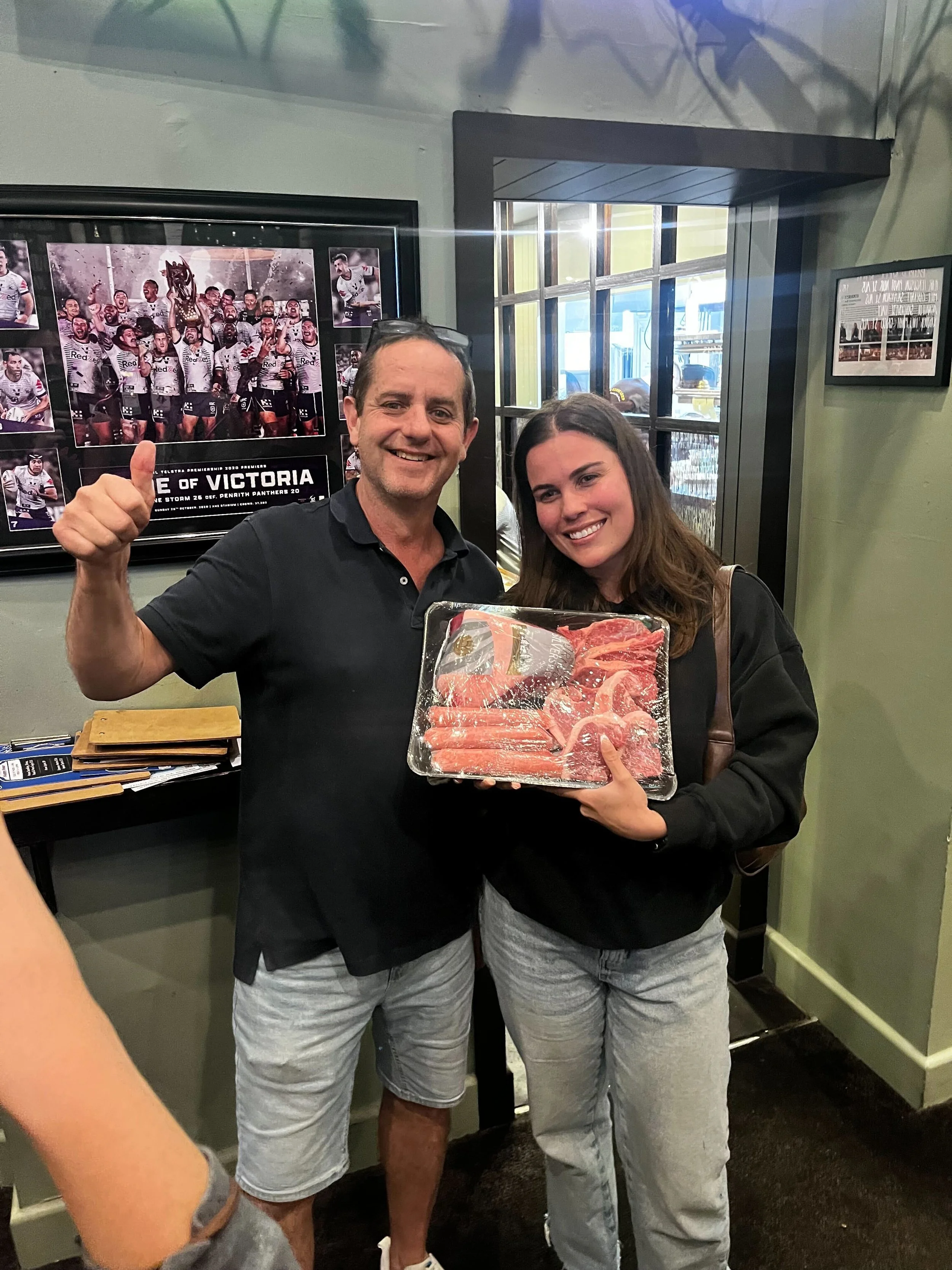 A man and a woman standing indoors, smiling at the camera. The man is giving a thumbs-up and holding a tray of raw meat. Behind them are framed photos of a sports team celebrating a victory.