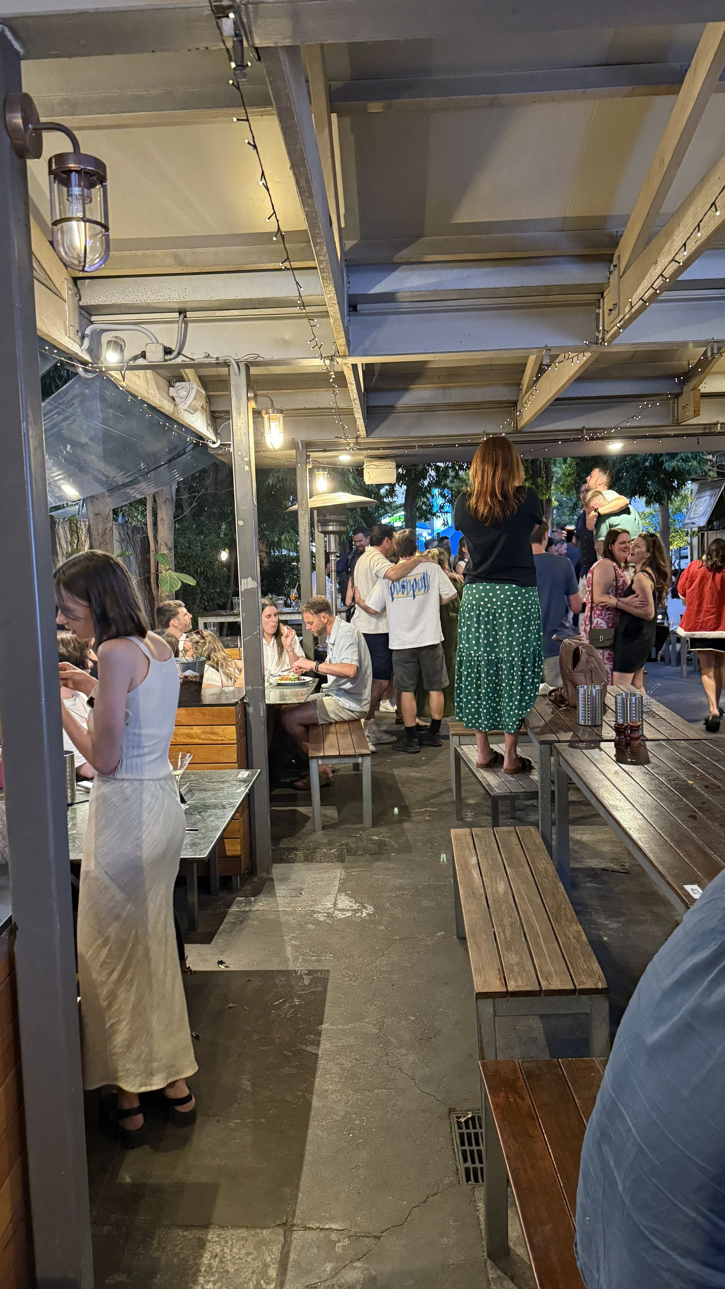 People socializing and dining in the beer garden with wooden tables, string lights, and a covered roof.