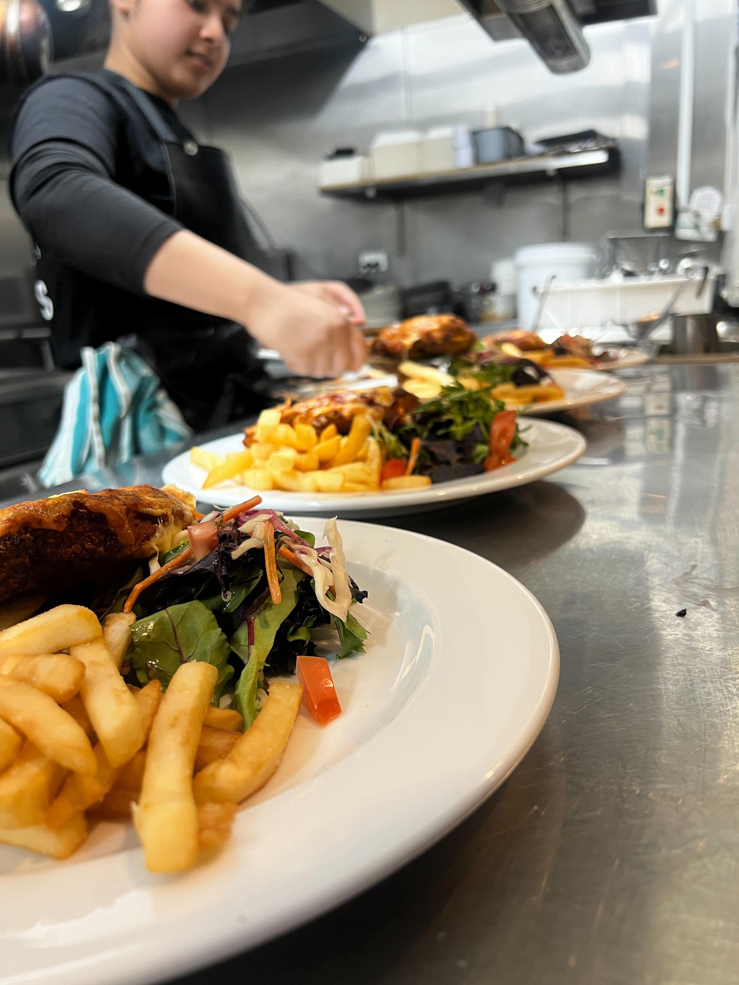 Close-up of a white plate filled with French fries, a fresh garden salad with lettuce, tomatoes, and shredded carrots, and a piece of cooked chicken, with a person preparing more food in a kitchen in the background.