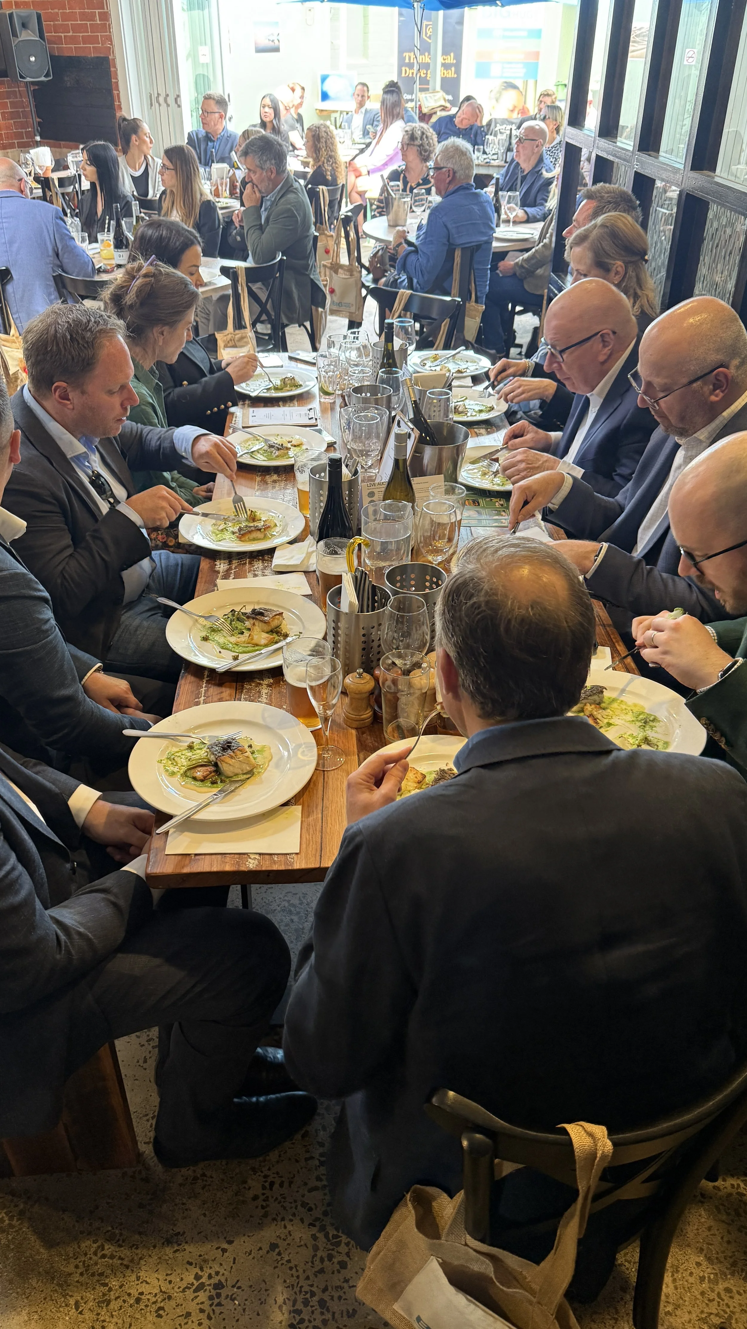 A group of people in business attire sitting together at a long wooden table in a restaurant, eating a meal. The table is set with white plates, glasses, and utensils, and the background shows more diners in a well-lit, modern dining space with large
