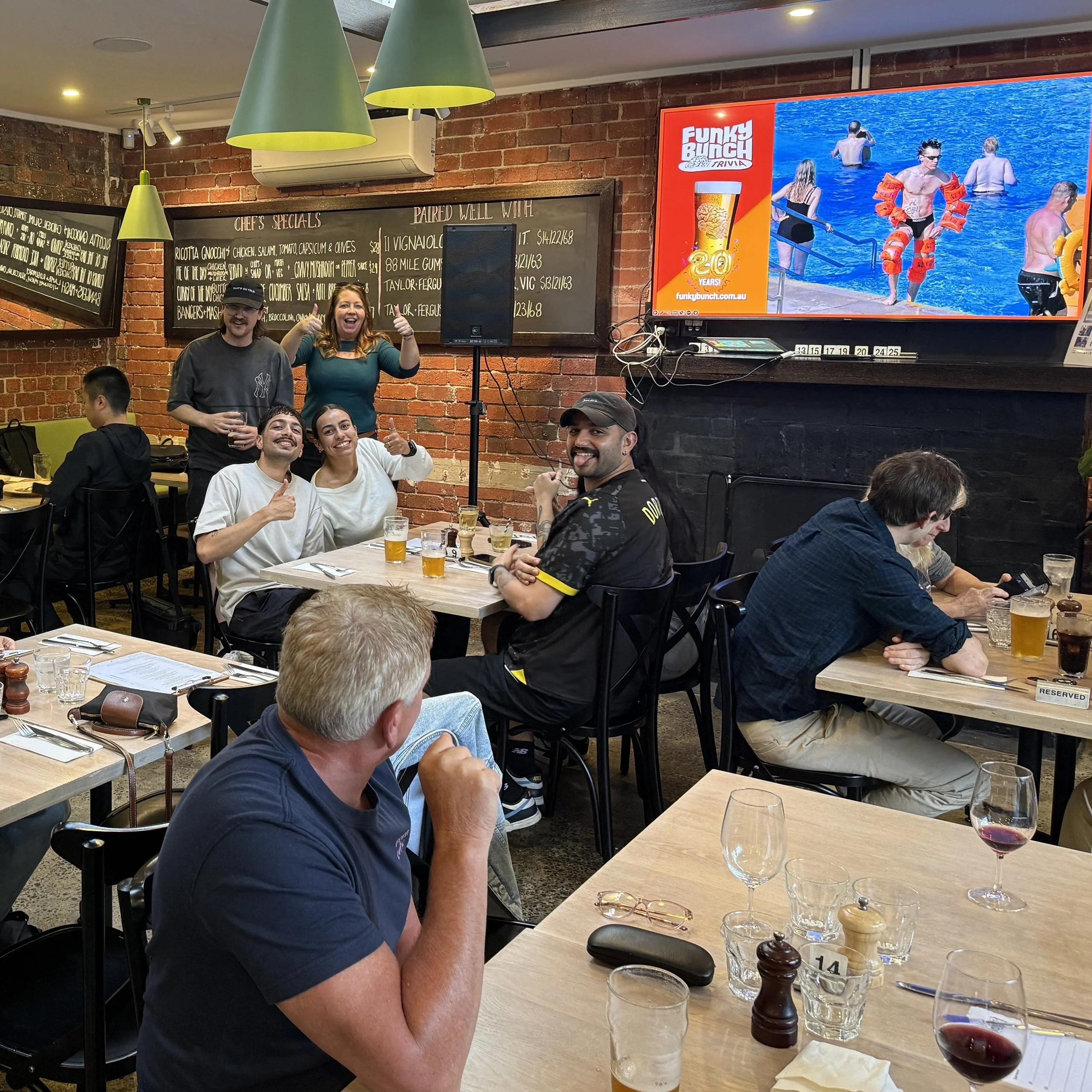 Group of people dining at a restaurant with brick walls and a large digital screen displaying an advertisement for Funky Bunch trivia.