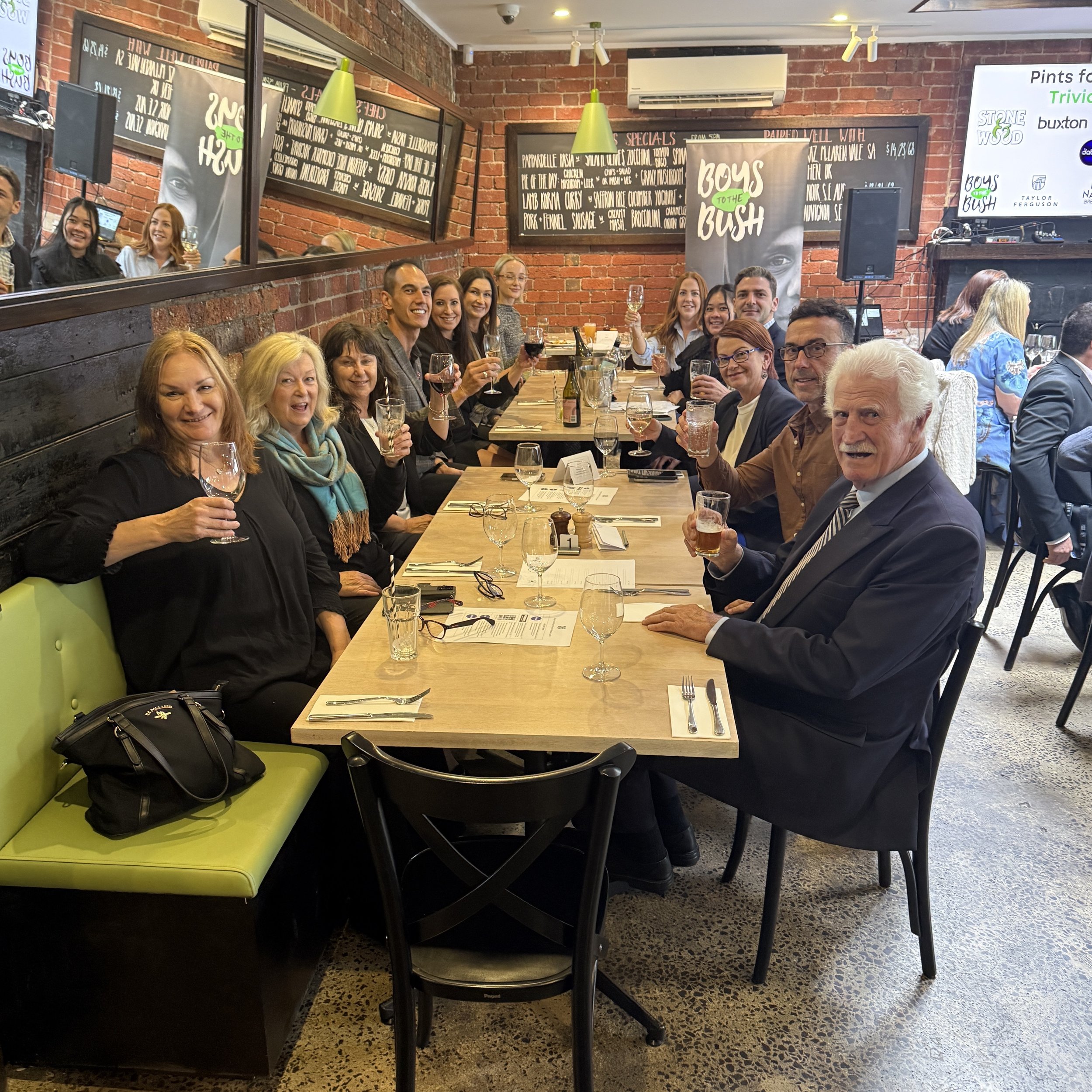 People sitting at a long dining table in a restaurant, smiling and holding drinks, with a brick wall and menu boards in the background.