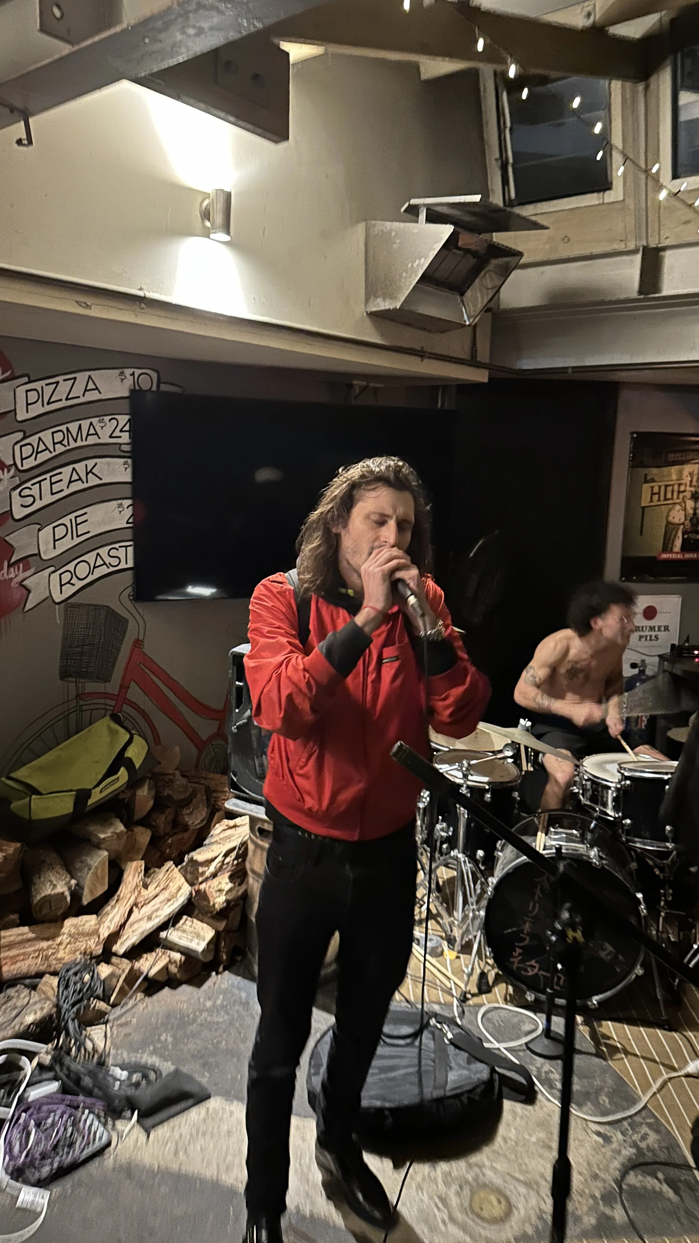A man with long hair singing into a microphone and another man playing drums behind him in the Palace Hotel beer garden at night.