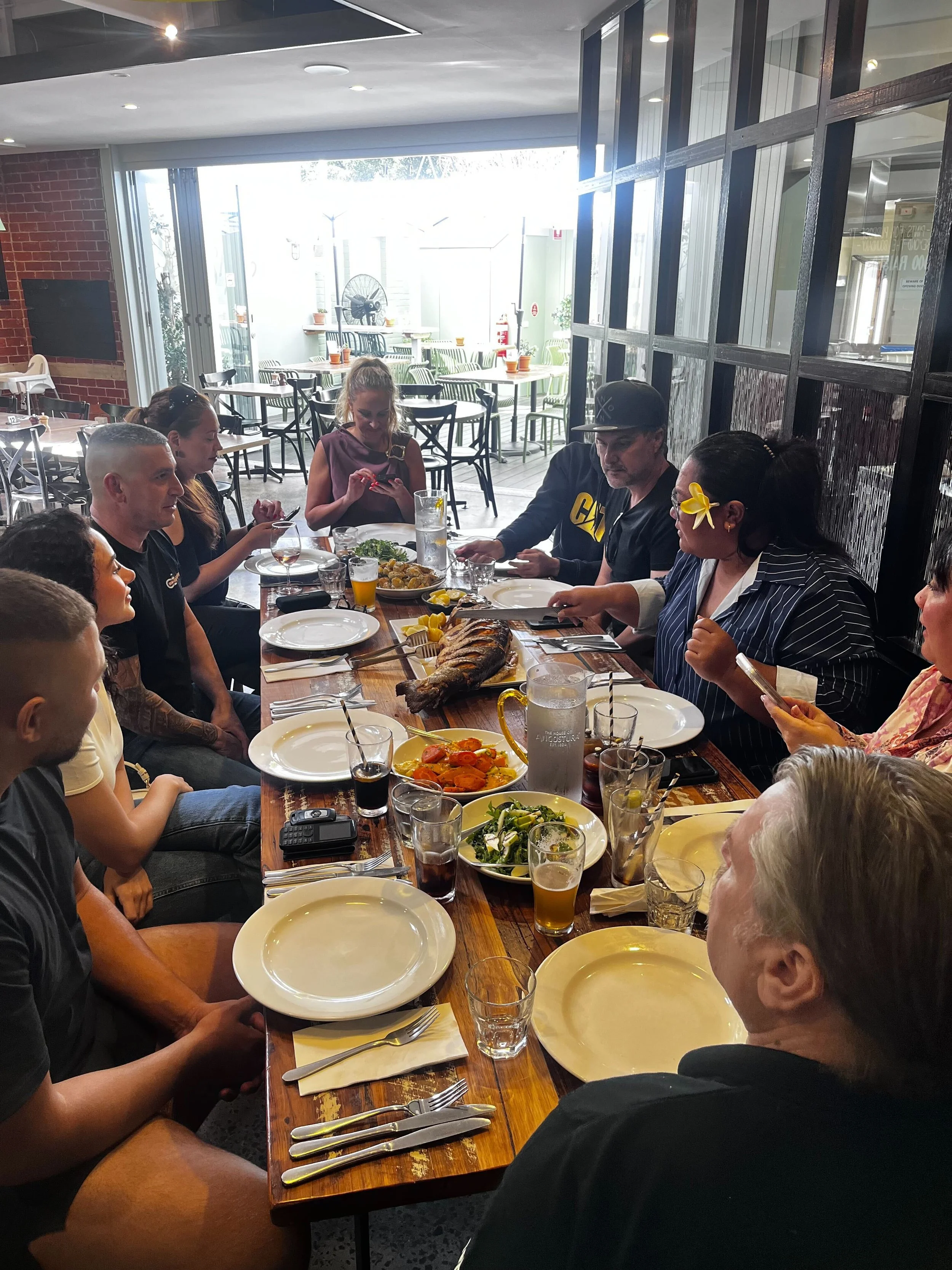 A group of people sitting around a long dining table in a restaurant, with dishes including a whole fish, salads, and drinks, during daytime with sunlight coming through the window.